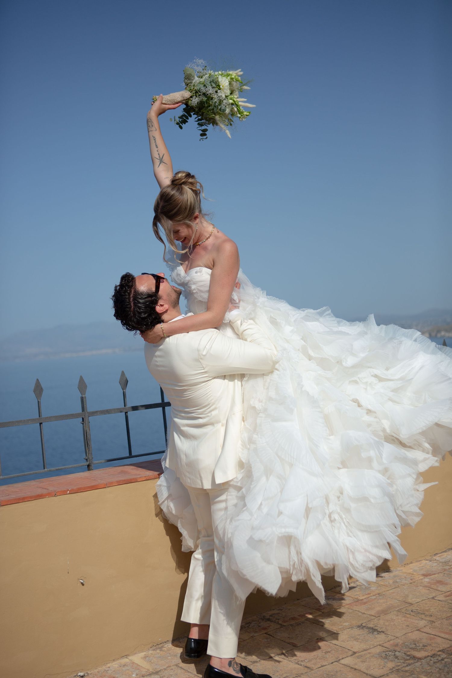 A couple in wedding attire dances and dips dramatically against a scenic ocean backdrop on a sunny day.