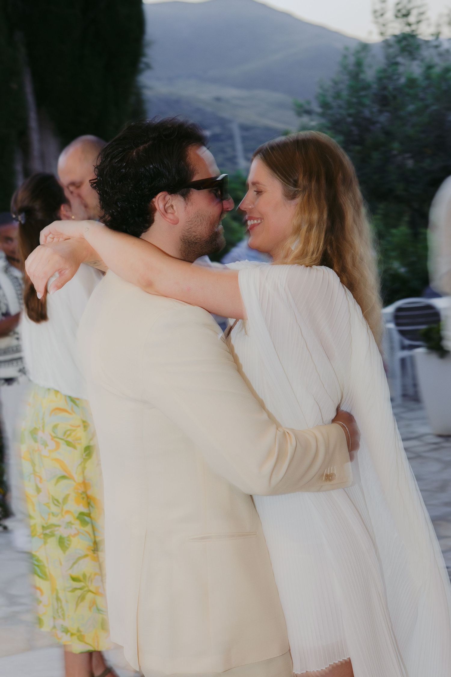 A couple in white outfits share an intimate dance moment outdoors with mountains visible in the background.