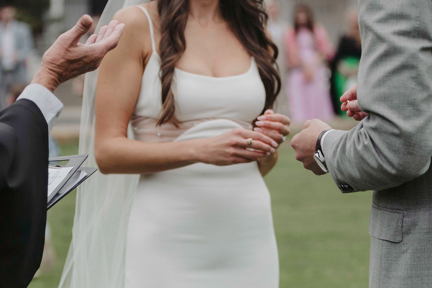 A couple stands close together at an outdoor wedding ceremony wearing a white dress and gray suit.