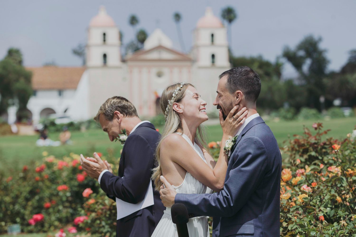 A romantic wedding moment captured in a garden setting with a church steeple visible in the background.