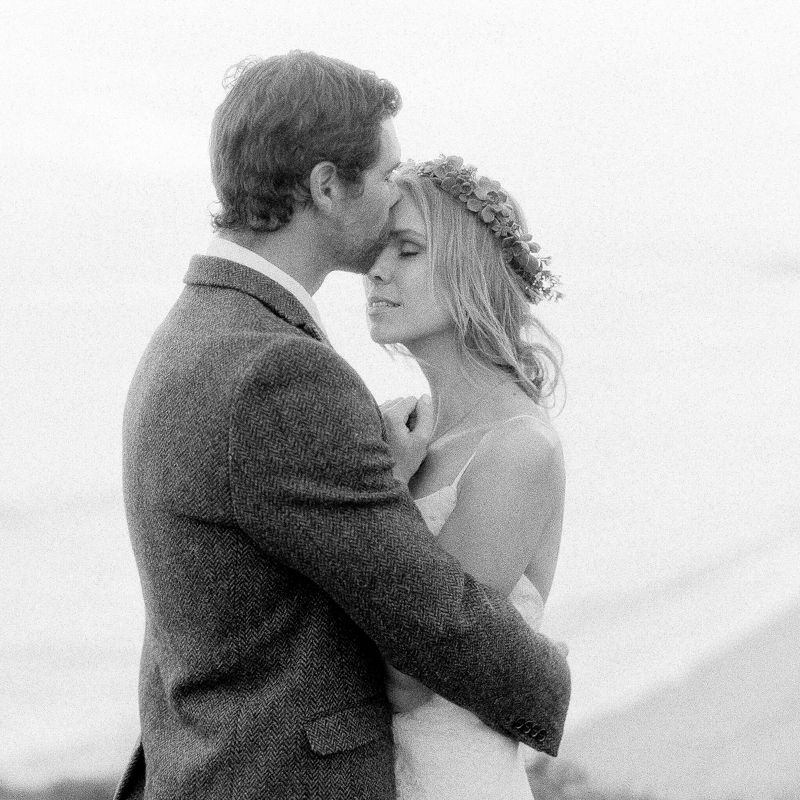 Black and white photo of a bride and groom embracing, bride wearing floral crown, romantic outdoor wedding portrait.