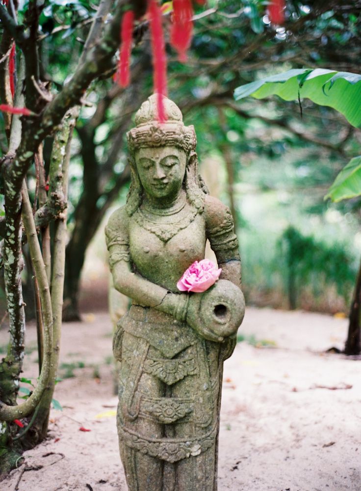 Ancient stone goddess statue holding a pink lotus flower in a lush tropical garden with red ribbons.