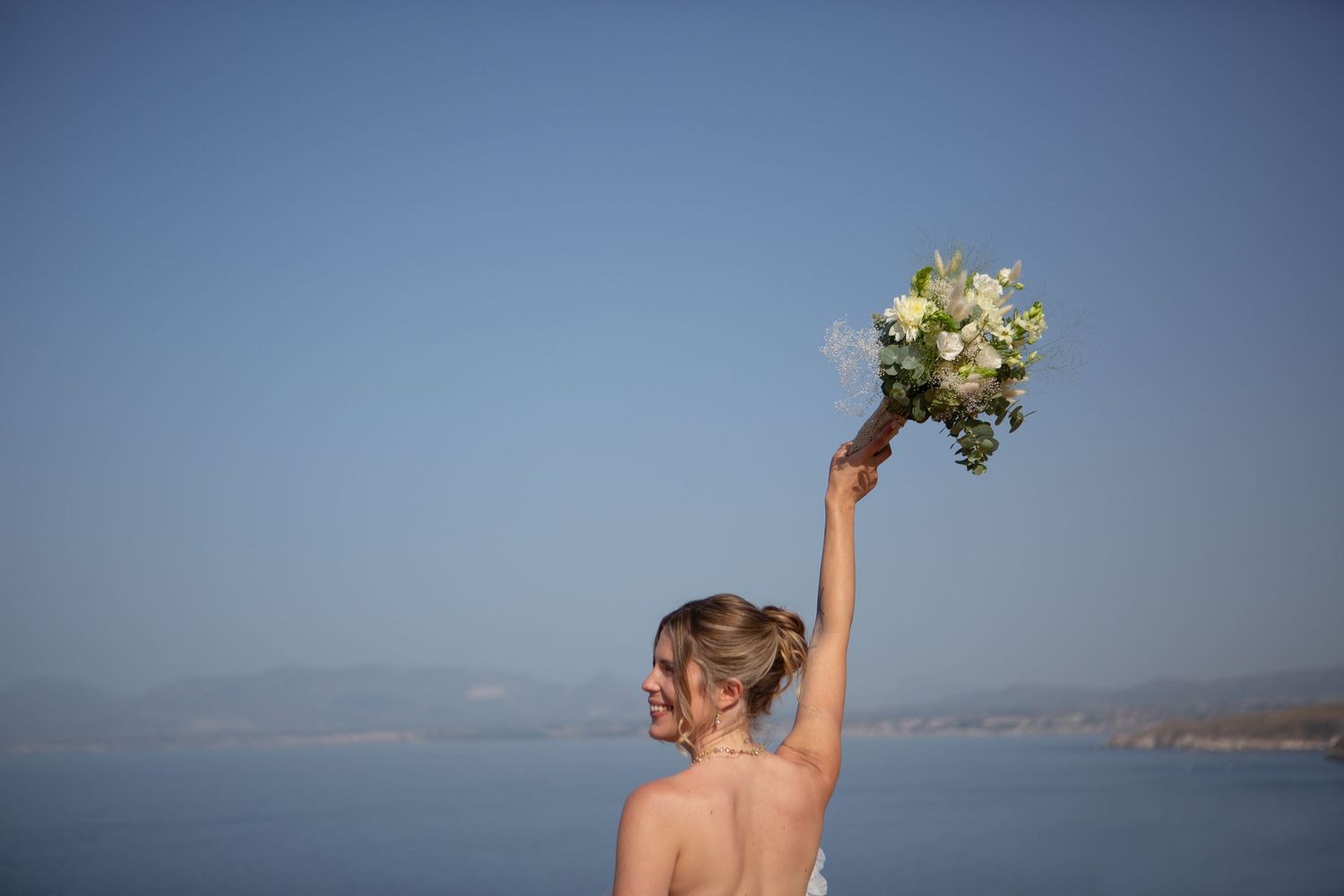 A bride in a white wedding dress holds her bouquet up against a blue ocean and sky backdrop.
