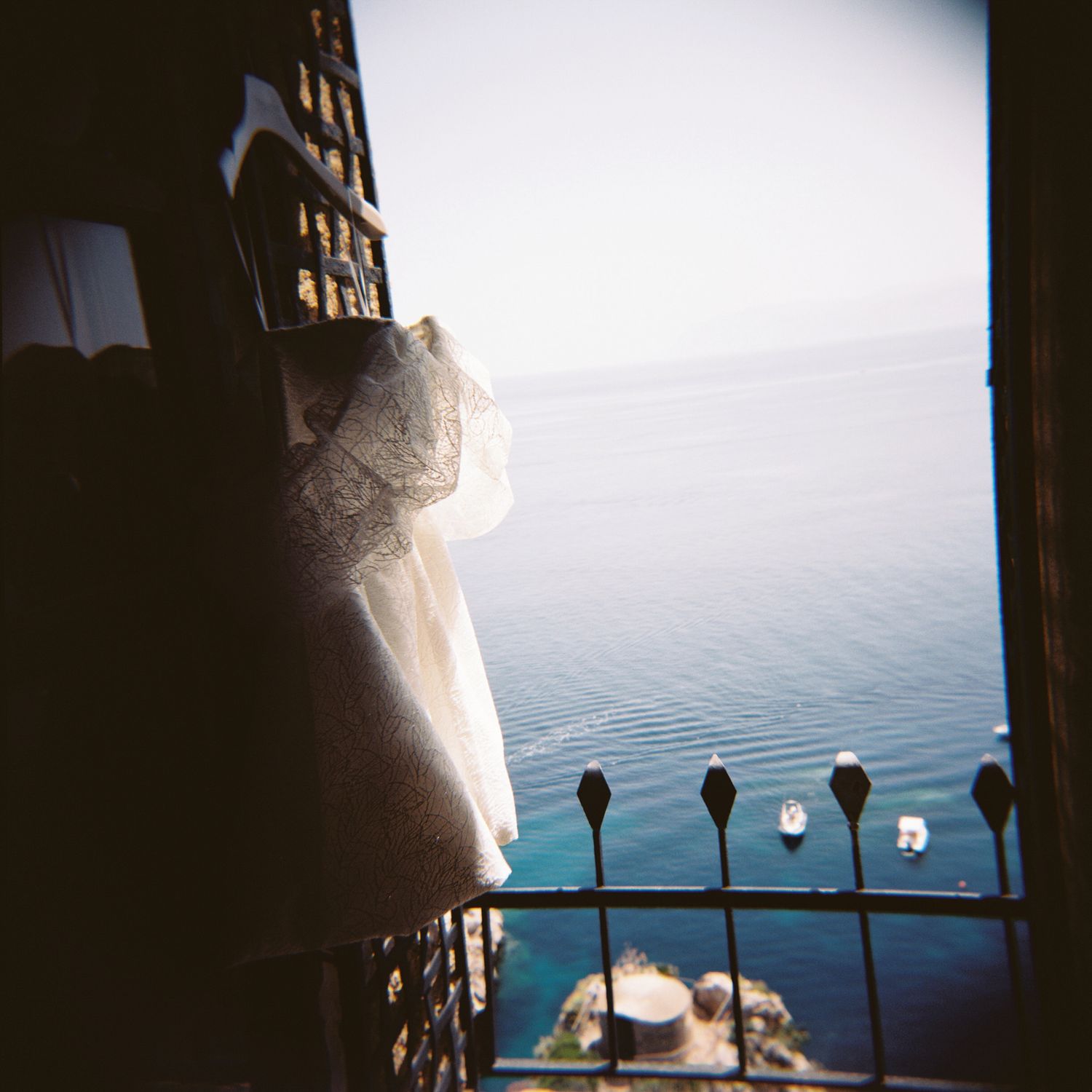 View of calm blue ocean waters from a balcony with decorative iron railings and white curtains billowing in the breeze.