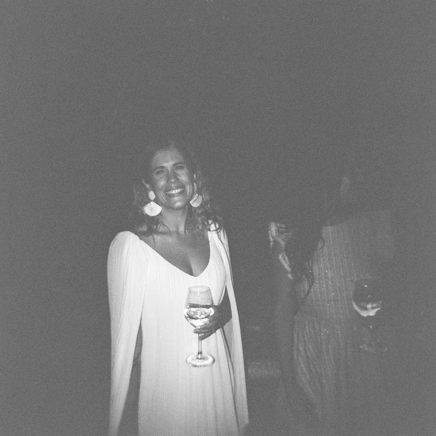 A person in a flowing white dress holds a wine glass while smiling at a nighttime gathering.