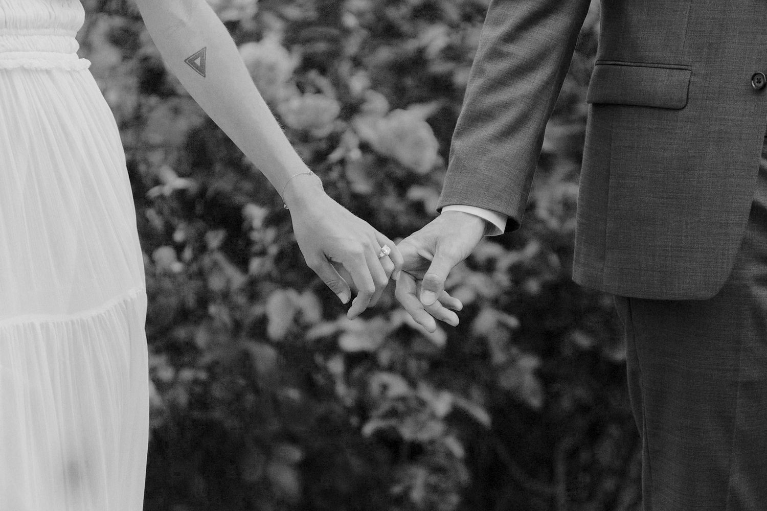 Black and white close-up photo of a couple holding hands with a blurred garden background.