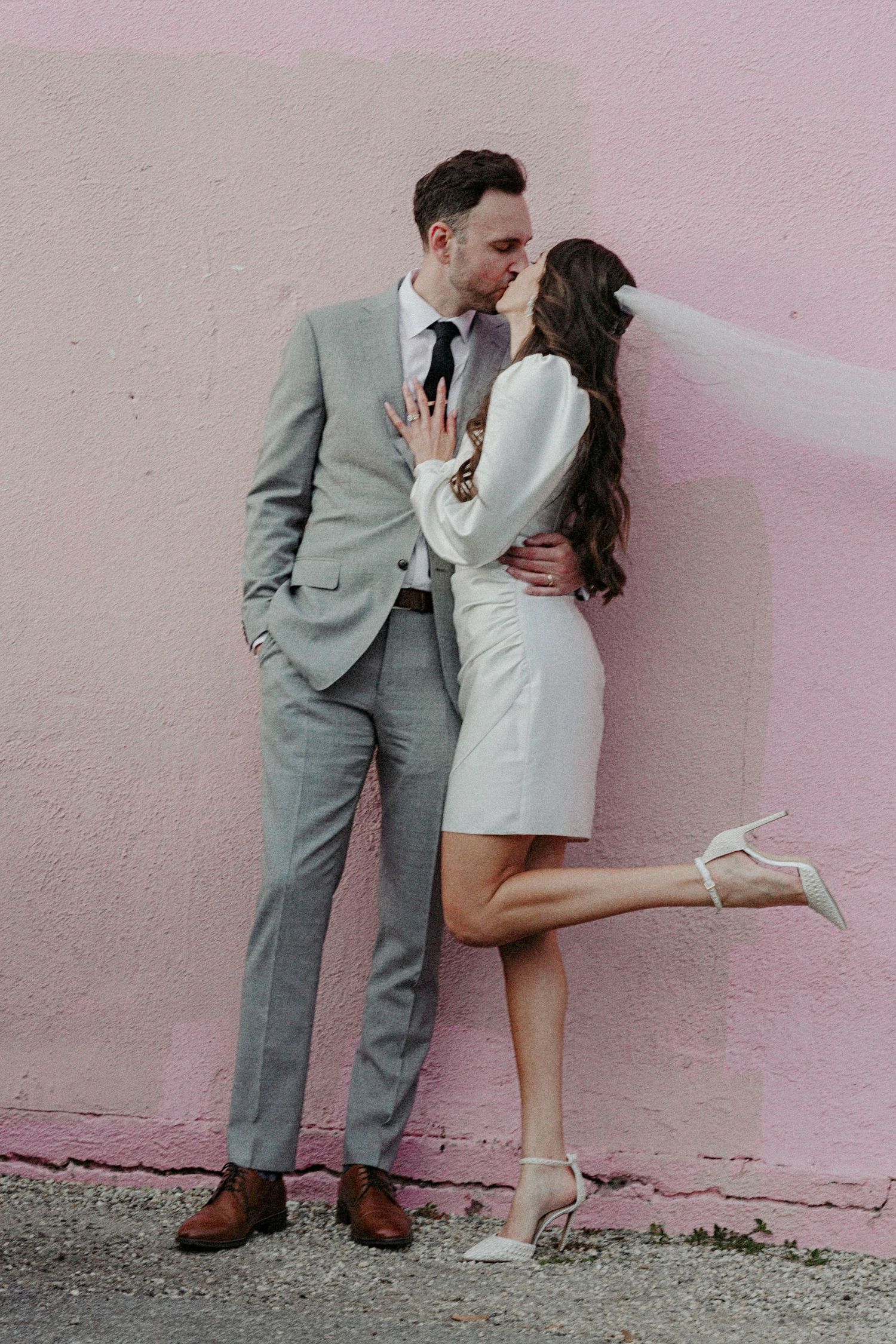A bride in a short white dress and groom in a gray suit share a romantic kiss against a pink wall.