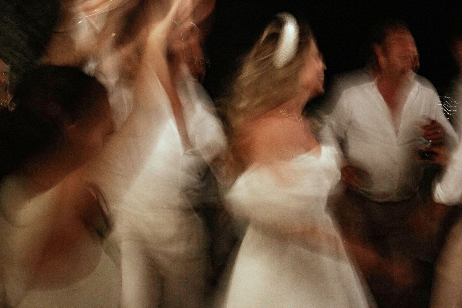Blurred motion of people dancing in white outfits at a lively celebration or wedding reception.