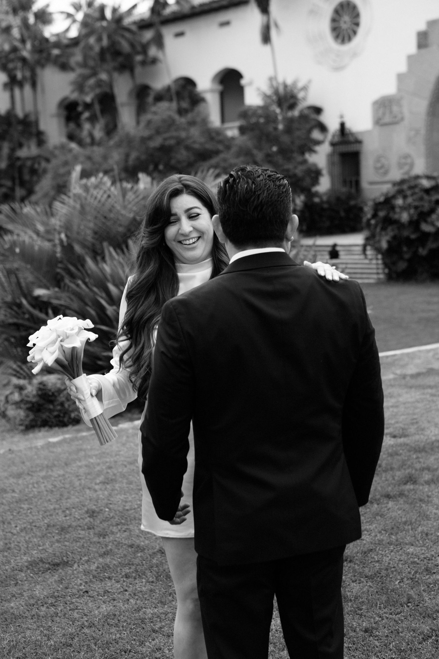 A couple in formal wedding attire walks together on a grassy lawn in front of a church in black and white.