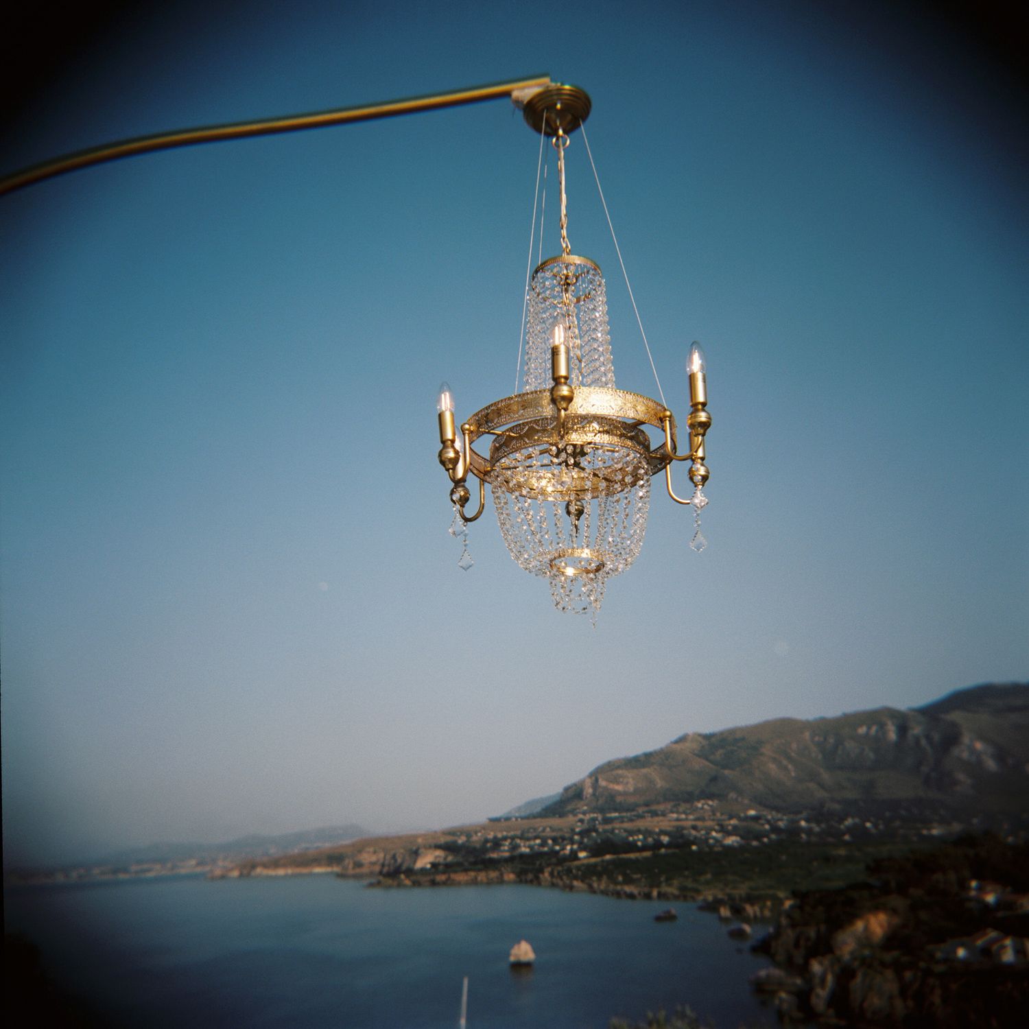 An ornate crystal chandelier hangs over a coastal mountain landscape with blue waters in the background.