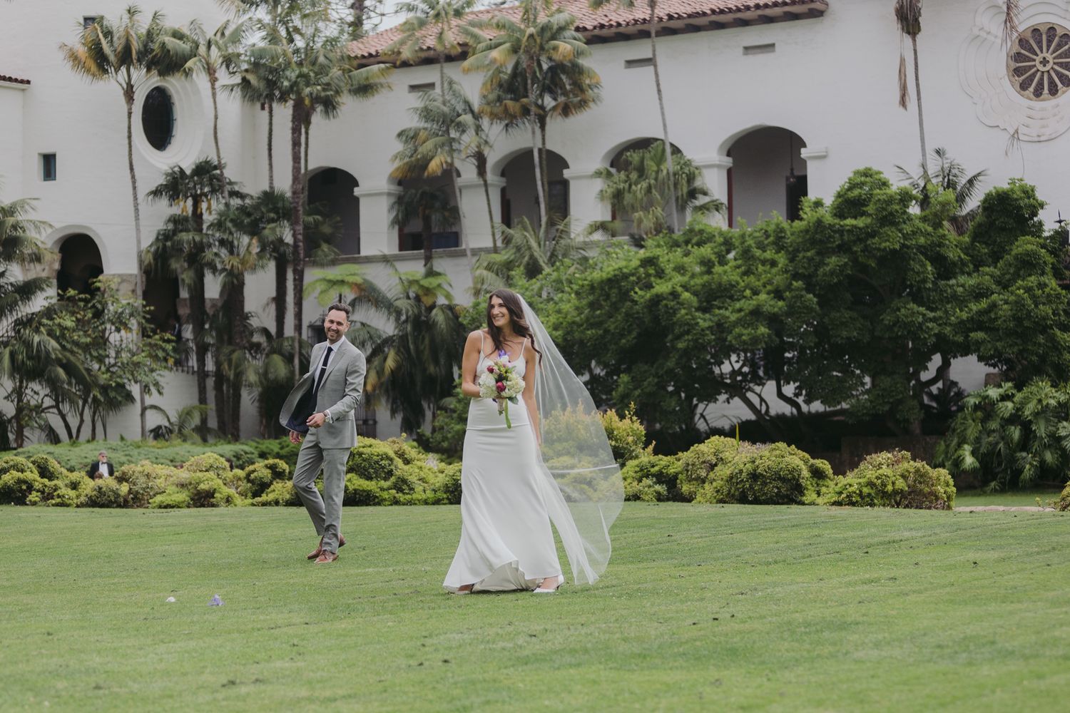 A couple walks together on a lush green lawn at a Spanish-style venue during a wedding celebration.