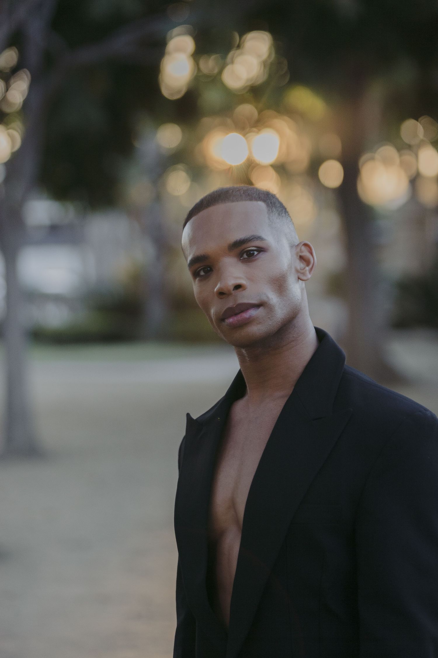Portrait photo of someone in a black blazer against a blurred outdoor background with bokeh lighting effects.
