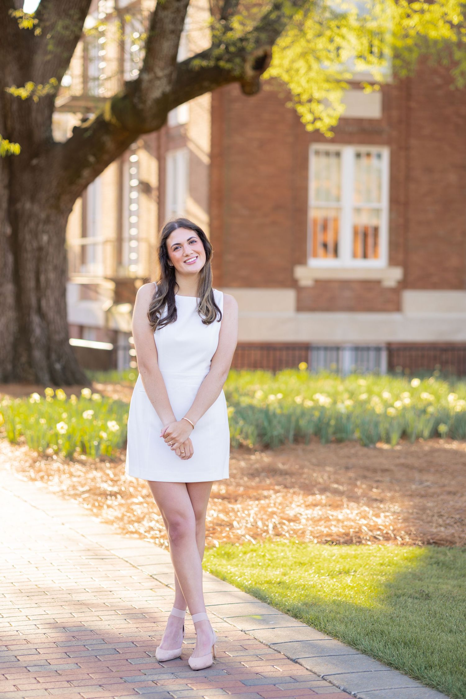 Young woman in white dress and heels smiling outdoors near brick building and blooming spring garden.