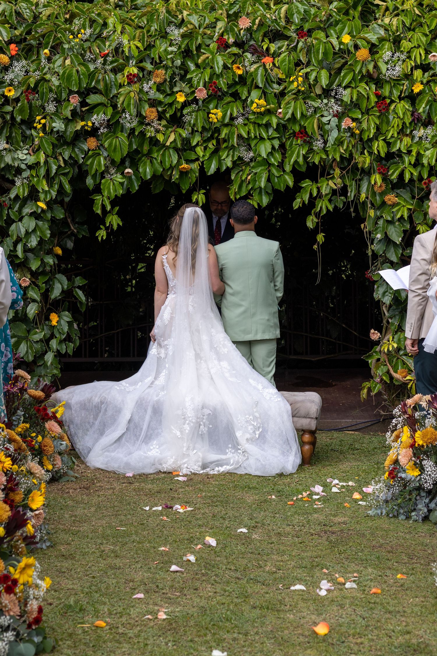 Wedding couple stands under floral arch with trailing bridal veil in garden setting.