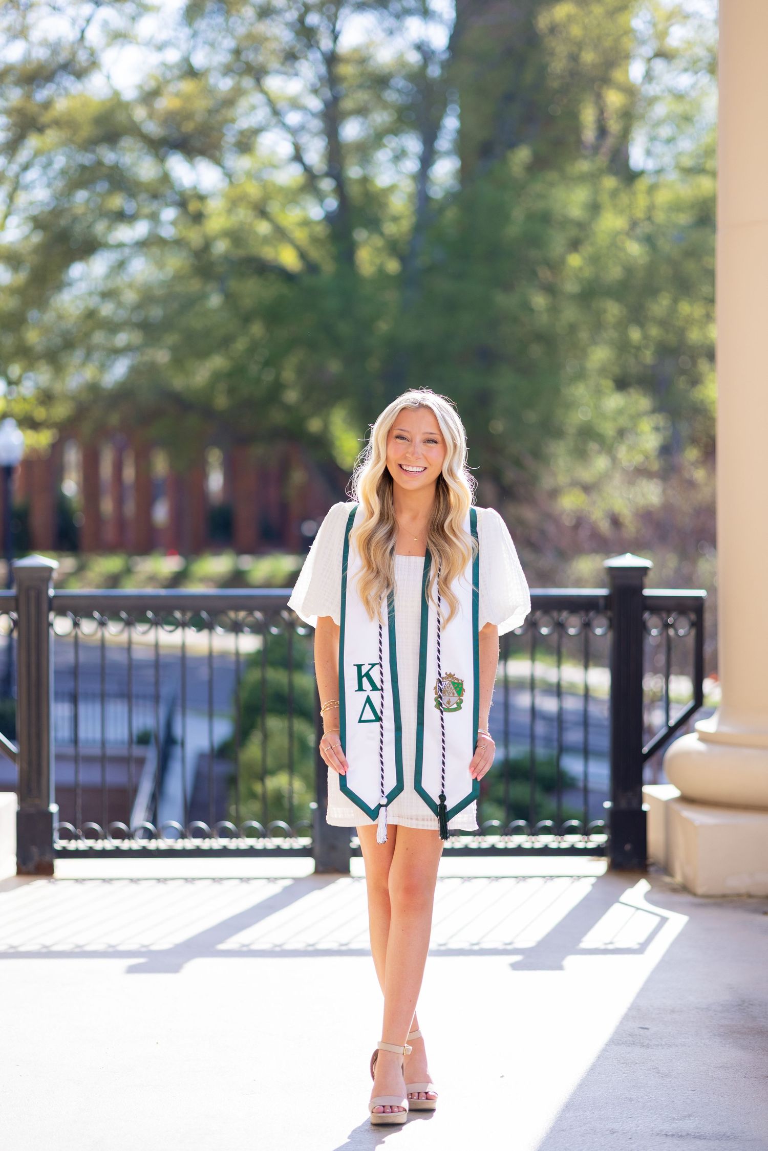 Smiling blonde woman wearing a Kappa Delta graduation stole poses outdoors on a sunny campus walkway.