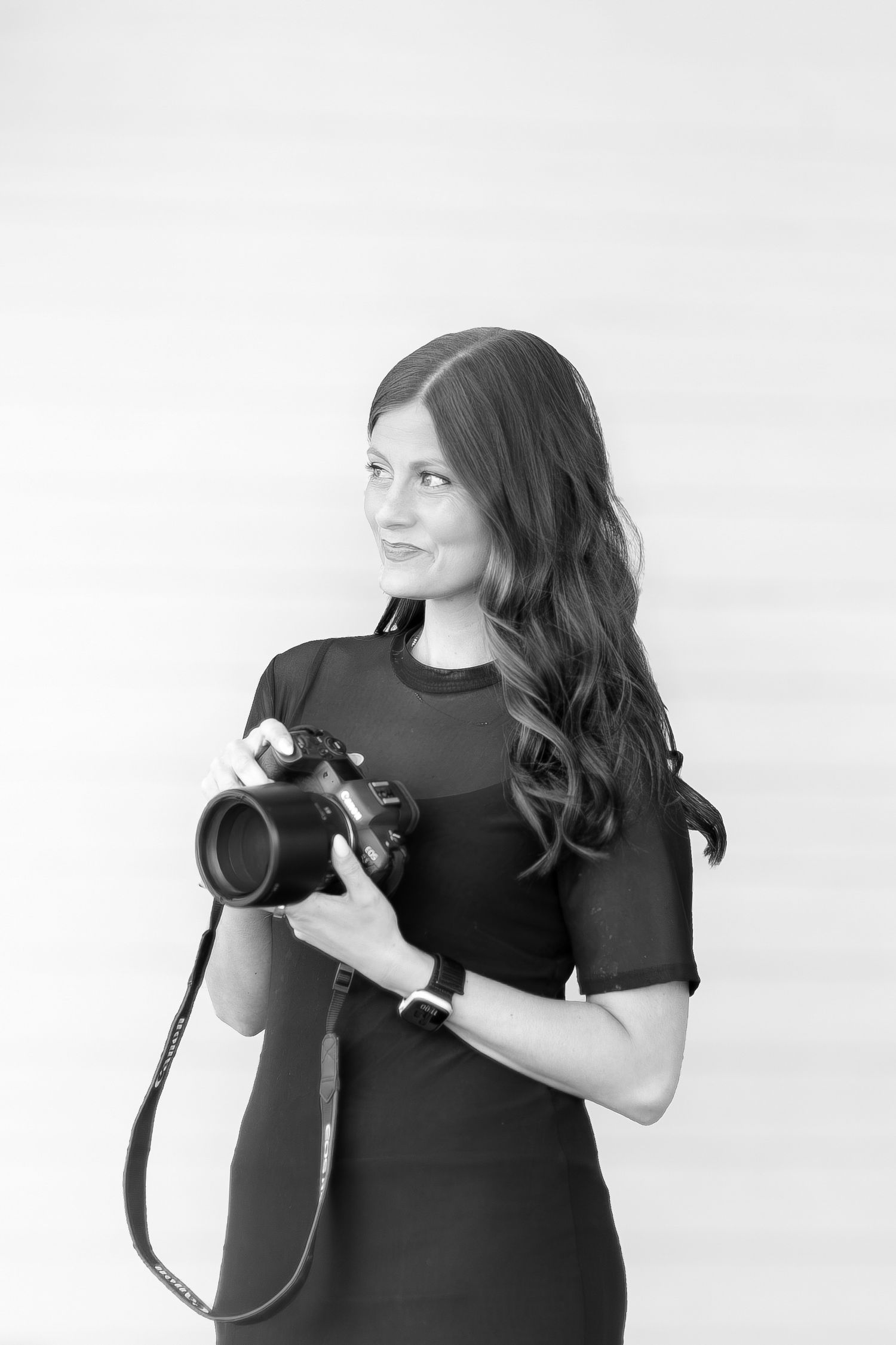 Black and white portrait of a smiling woman with long hair holding a DSLR camera, wearing a dark dress.