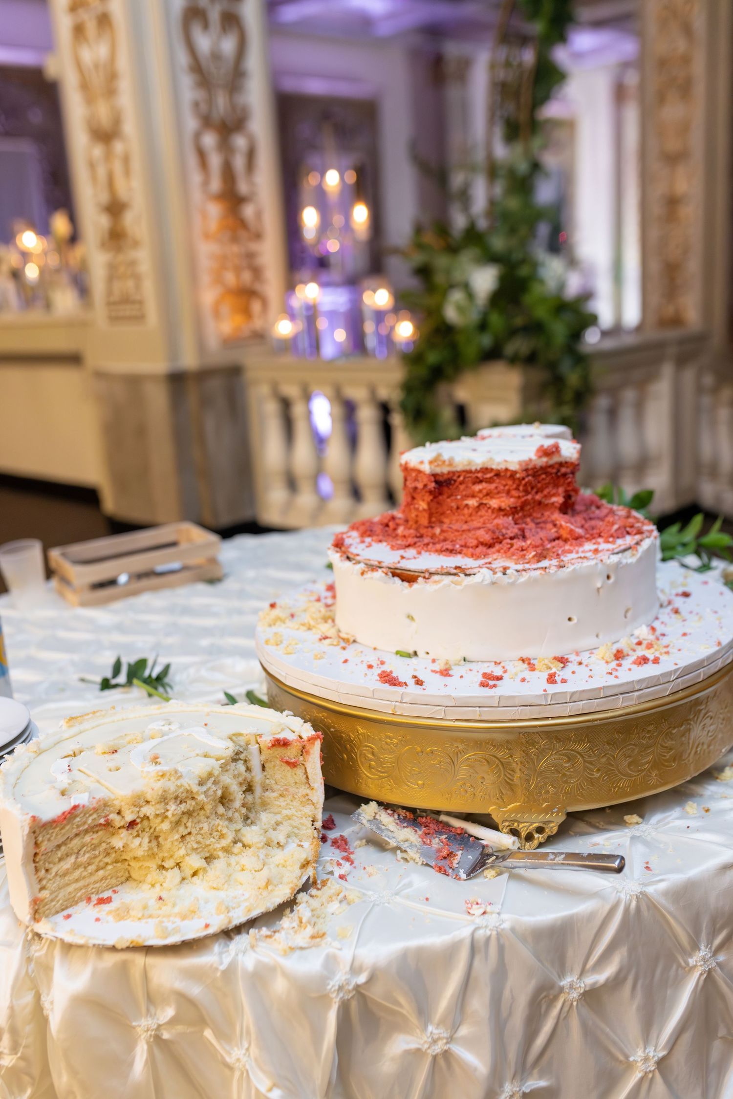 Two-tier white wedding cake with red velvet interior, partially cut, displayed on gold stand at elegant reception venue.