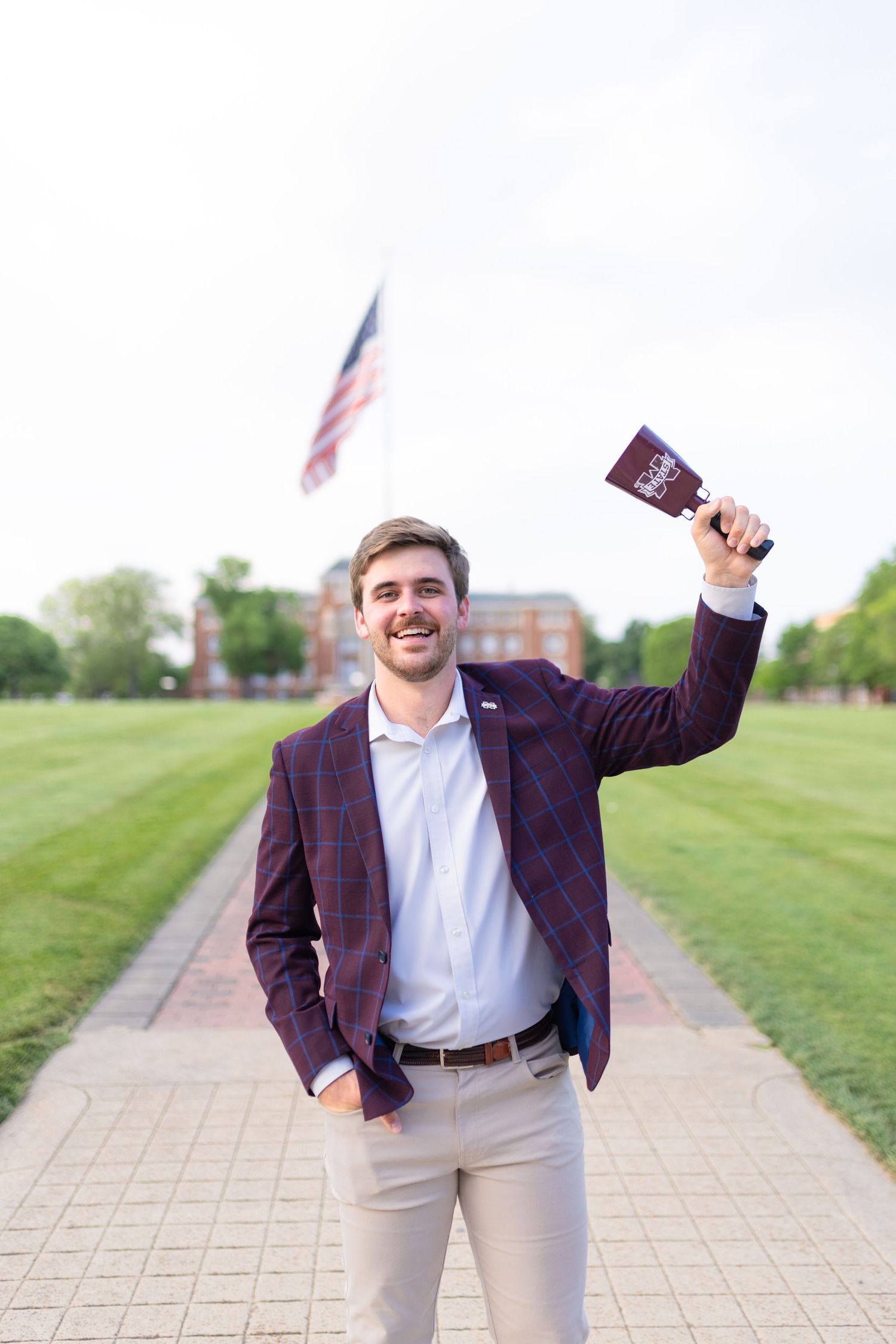 Graduate in plaid blazer celebrates with diploma on campus walkway.