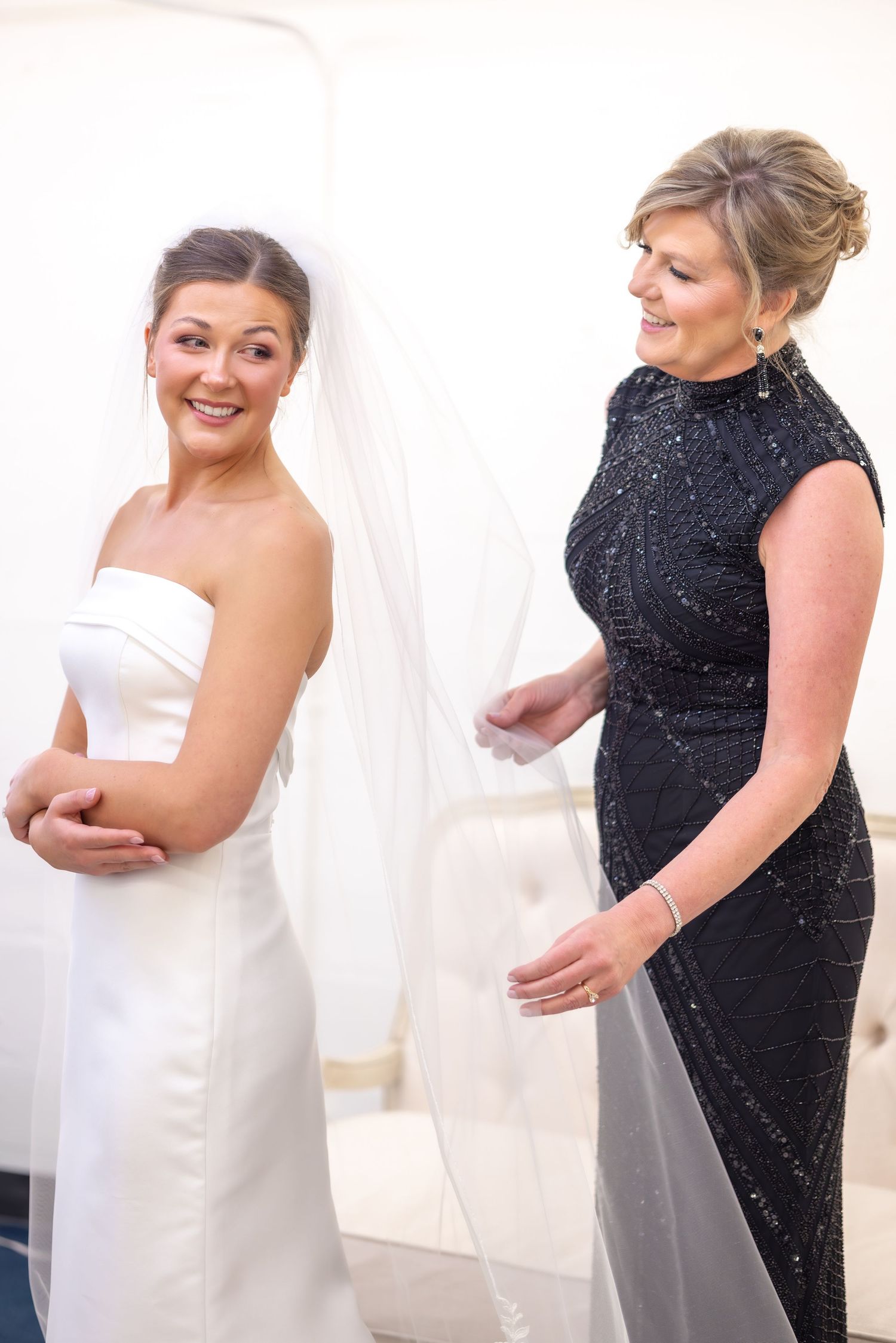 Bride in white strapless gown and veil smiles as woman in black beaded dress adjusts her veil before wedding ceremony.