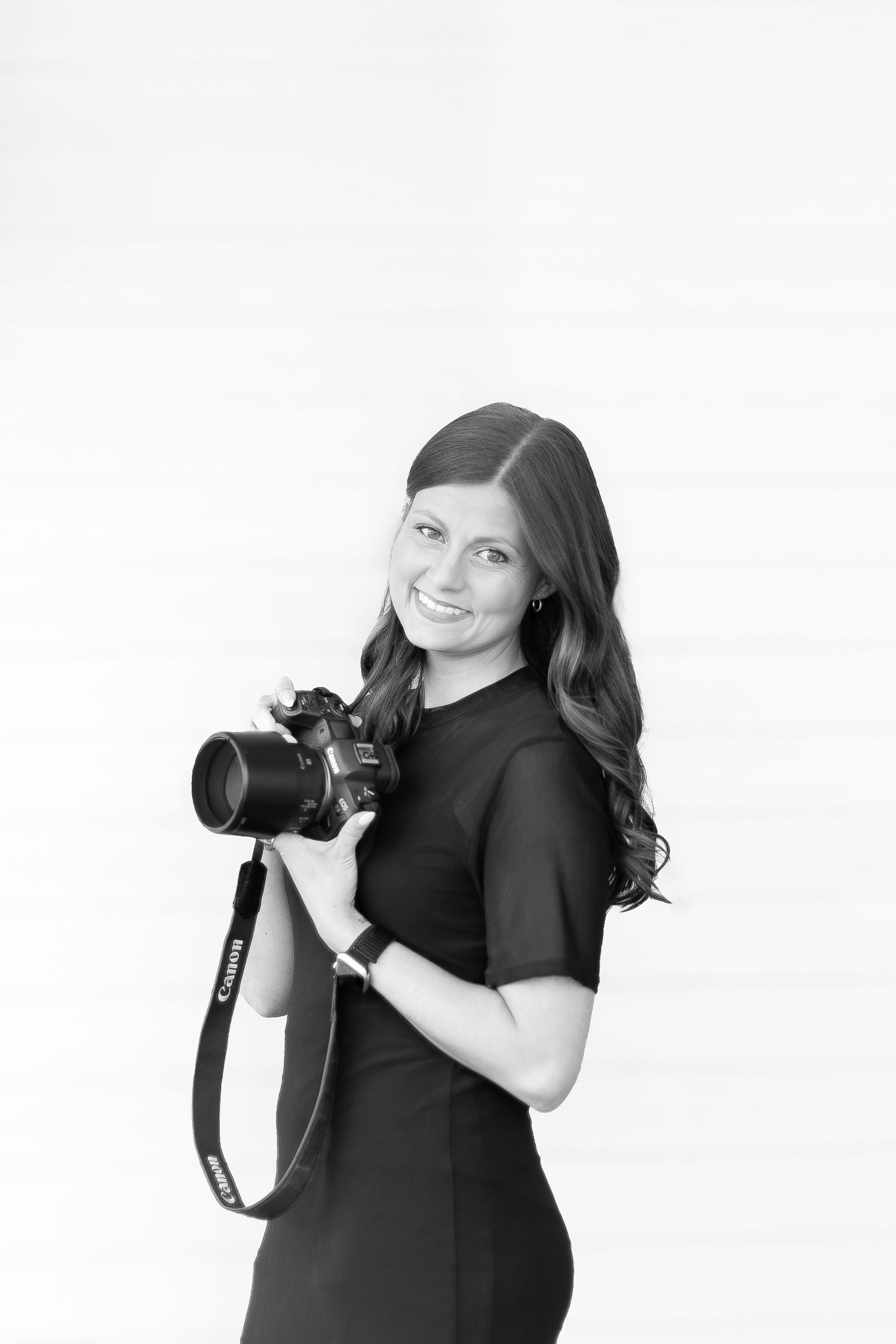 Black and white portrait of a smiling woman holding a Canon camera, wearing a black dress, on white background.