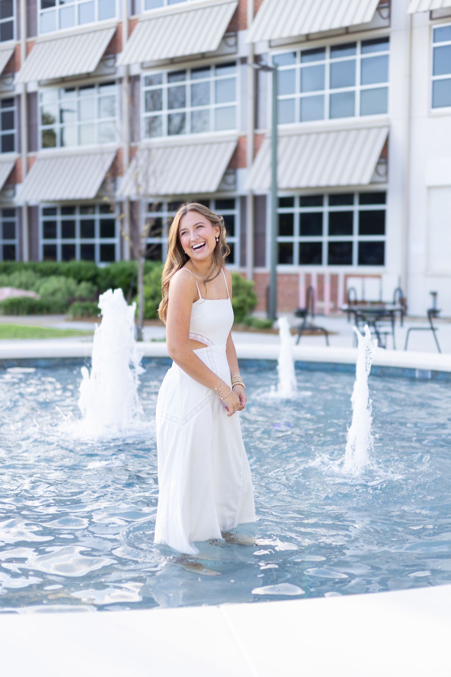 A person in a white dress poses by a decorative water fountain on a sunny day.