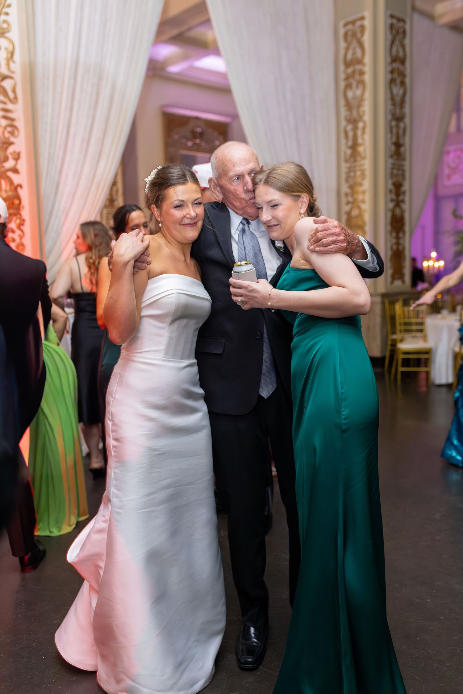 Bride in white gown hugs elderly man and woman in green dress at elegant wedding reception.