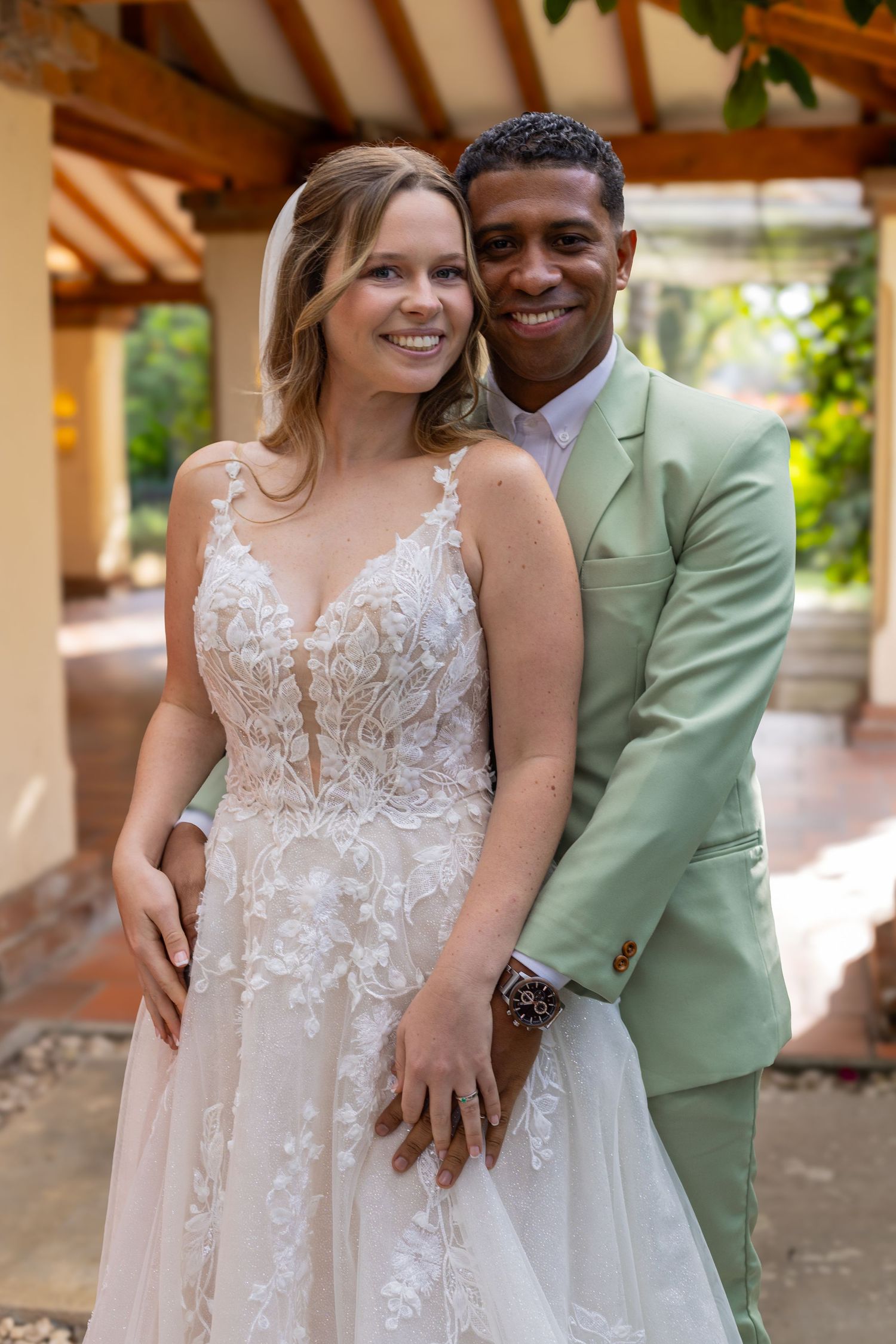 A smiling couple in wedding attire, bride in lace gown and groom in mint suit, posing outdoors.