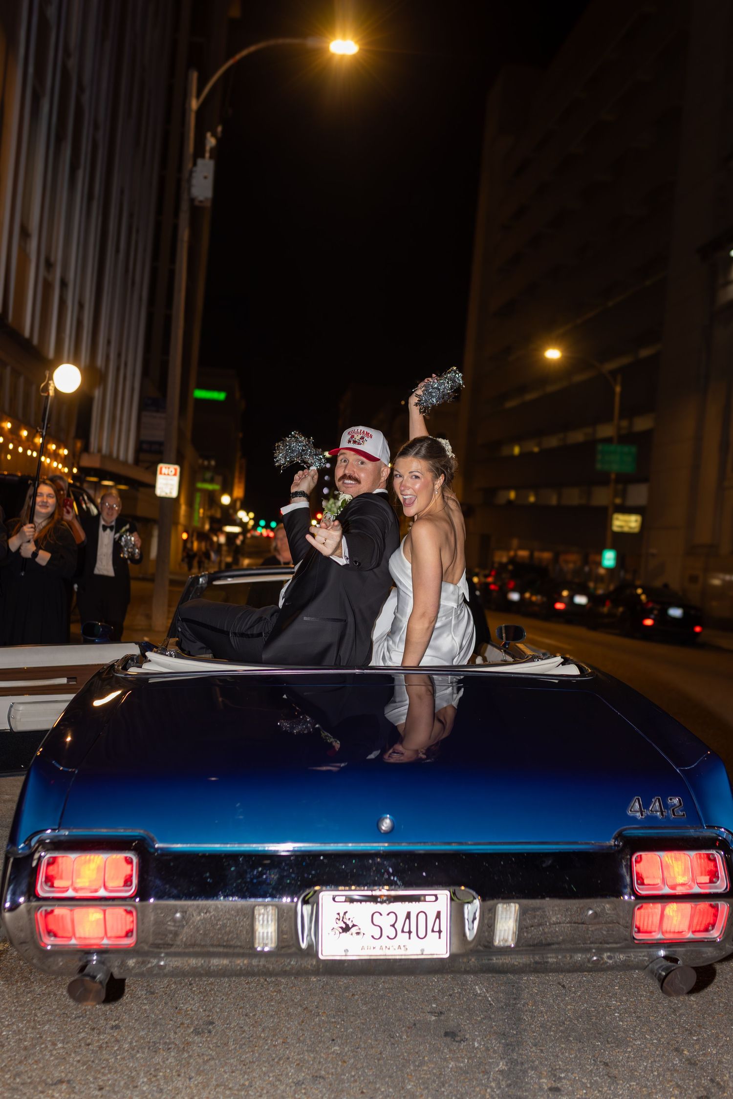 Couple celebrating in blue classic convertible car at night on city street with pom-poms.
