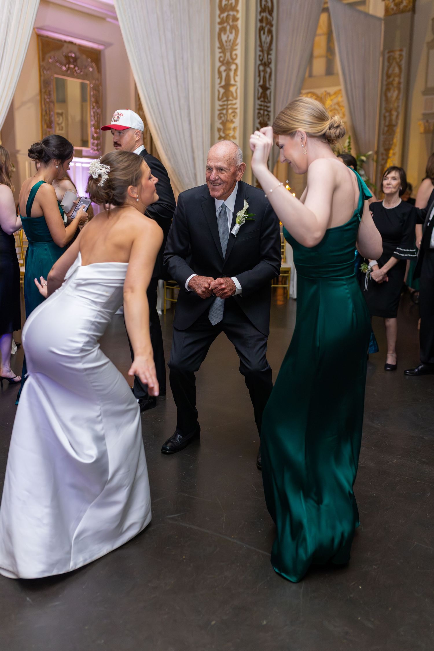 Bride and bridesmaid dancing joyfully with older man at elegant wedding reception in ornate ballroom.