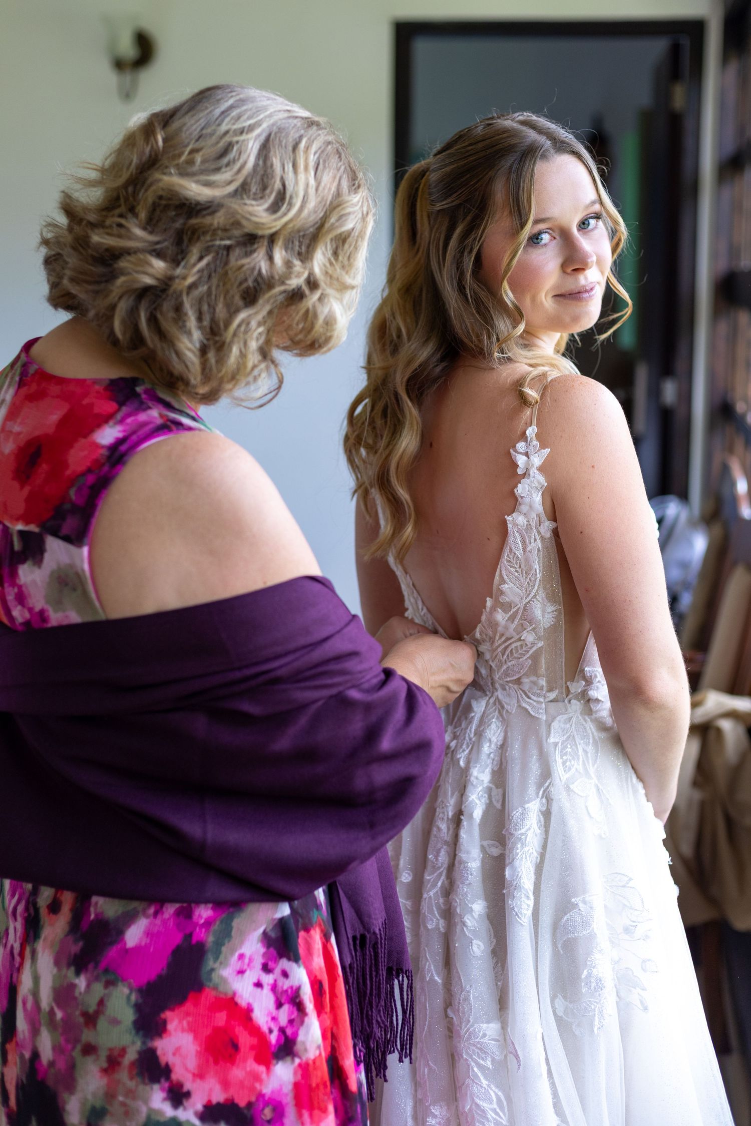 Bride in backless lace wedding gown being helped by woman in floral dress with purple shawl.