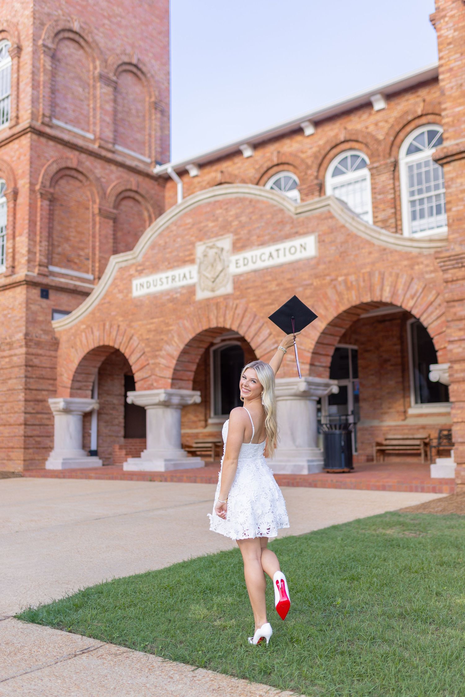 Graduate in white dress poses with diploma in front of historic brick university building.