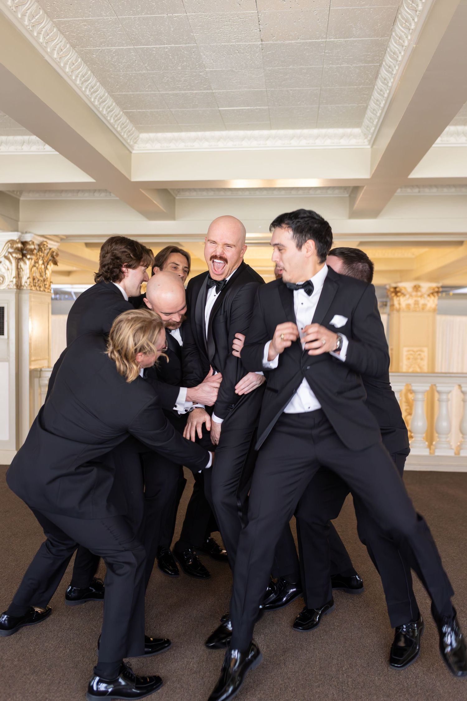 Groomsmen in black tuxedos playfully surround laughing bald groom in elegant wedding venue ballroom.