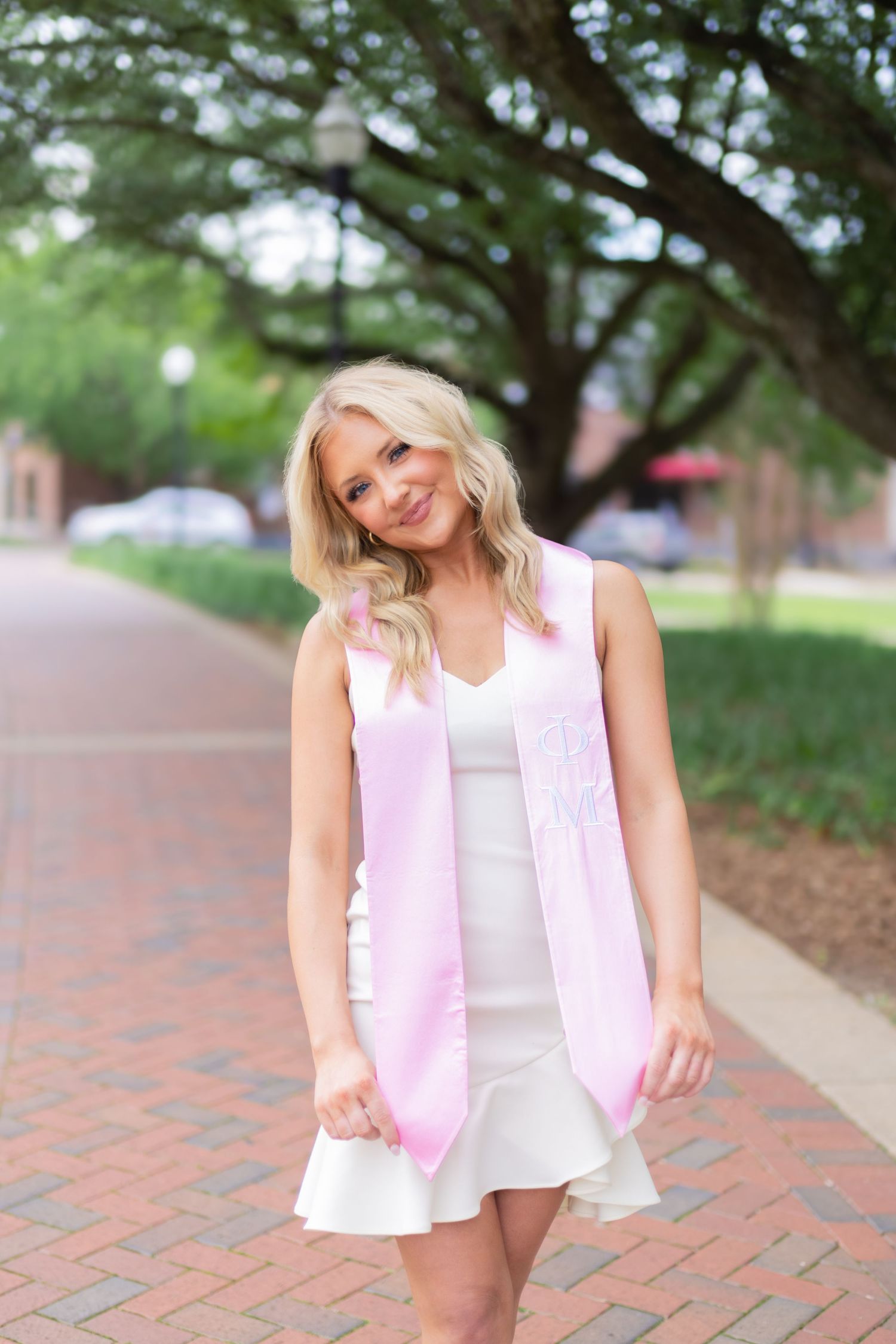 Woman in white top and pink vest poses playfully on brick walkway outdoors.