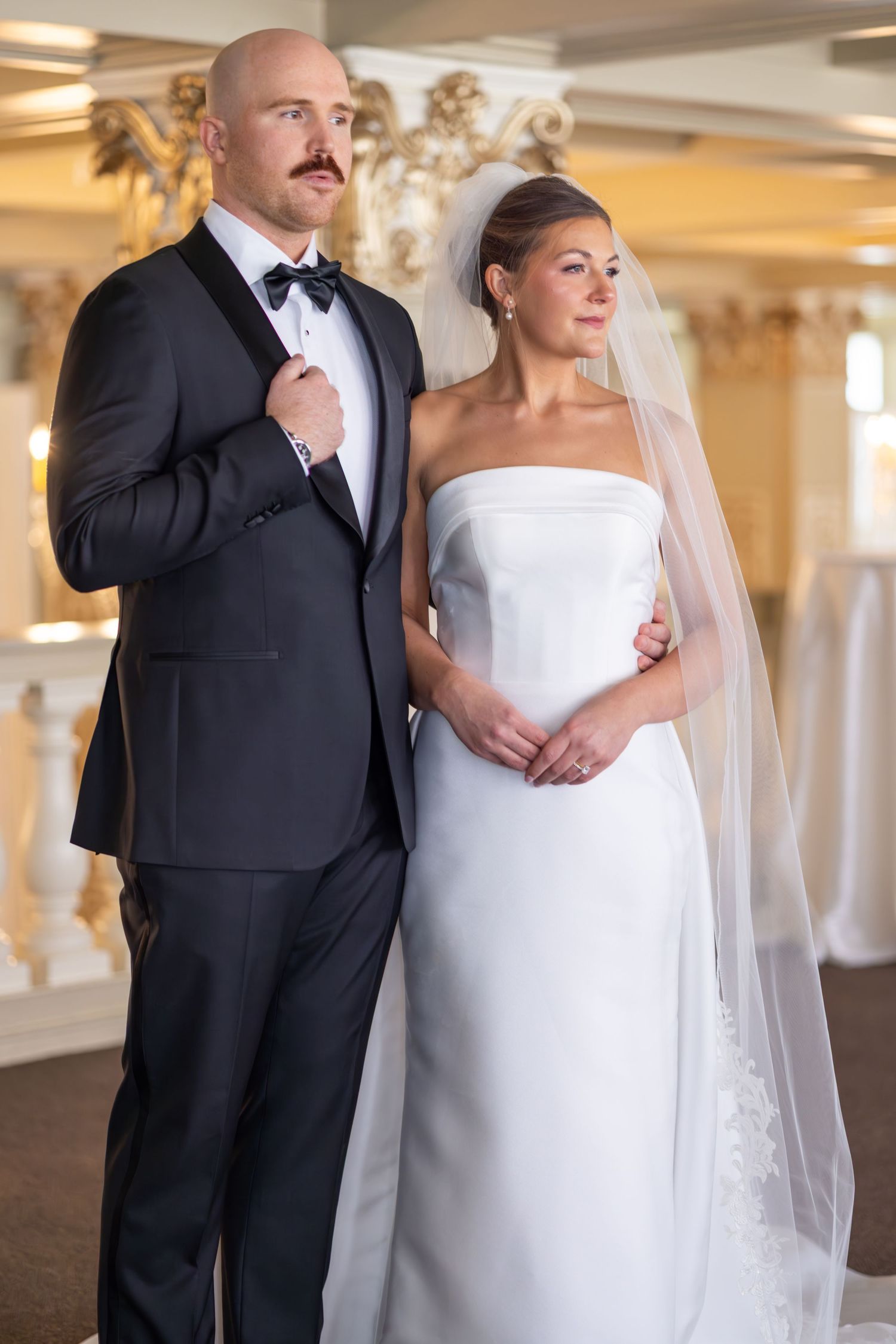 Elegant bride and groom posing together in a ballroom, wearing a white strapless gown with veil and black tuxedo.