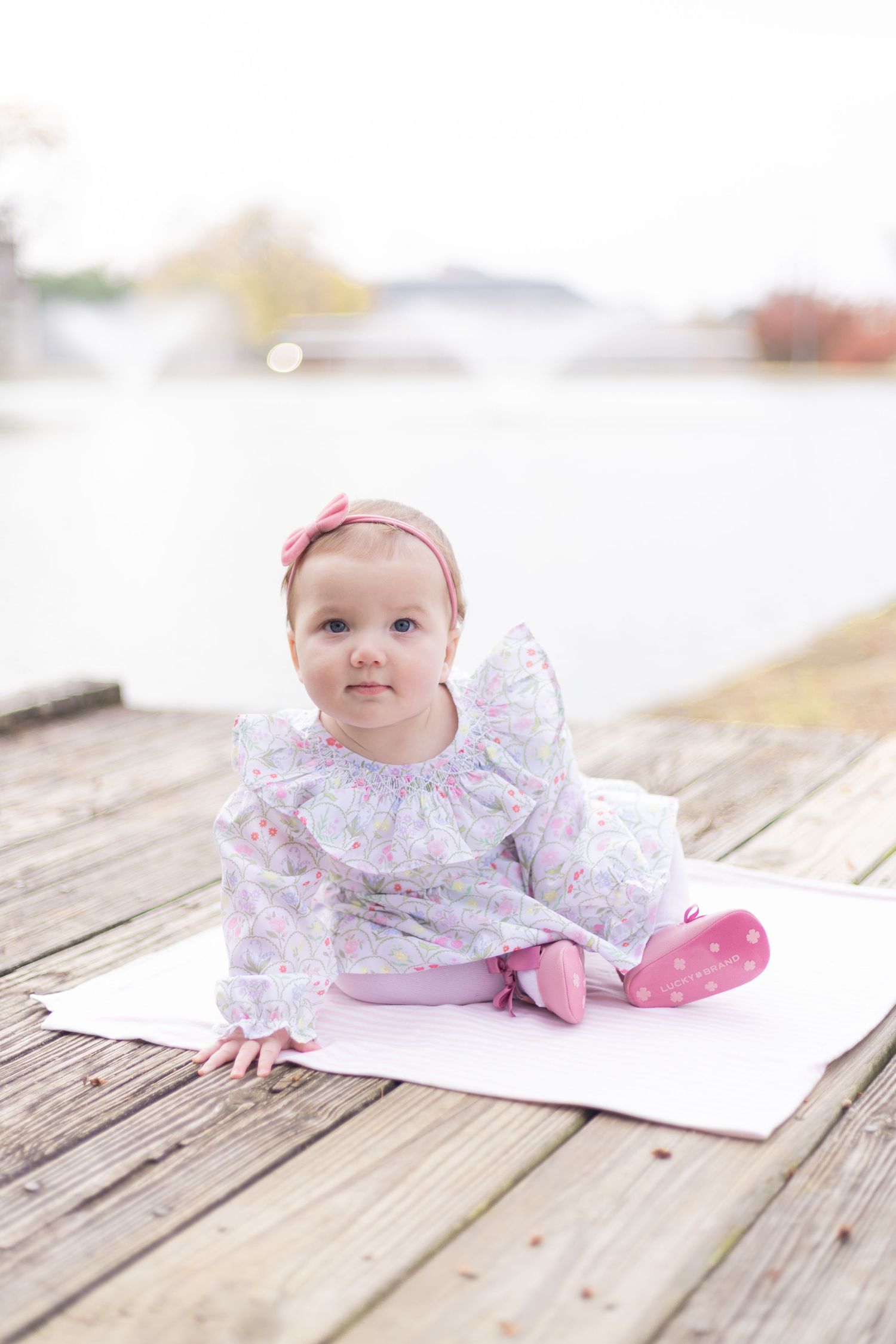 Baby in floral dress with pink bow lying on wooden deck during photo session.