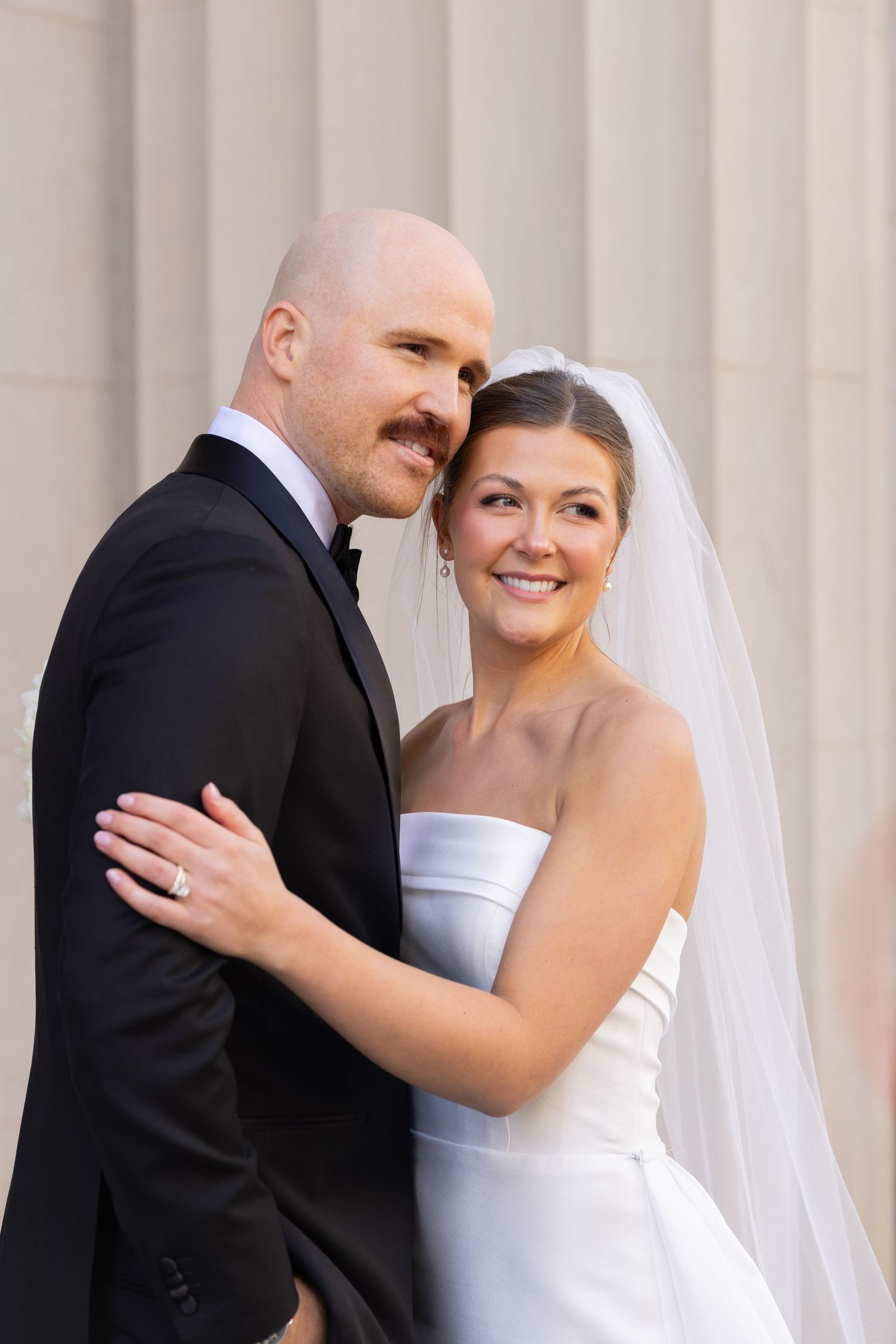 Bride and groom embracing outdoors, she wears a strapless white gown with veil, he wears a black tuxedo.