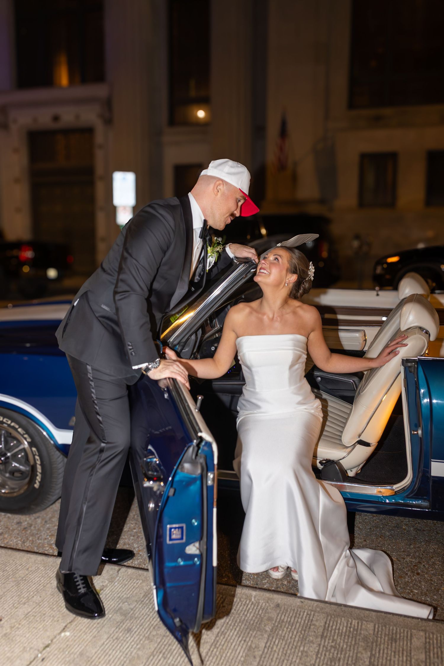 Bride in white strapless gown and groom in black suit share a romantic moment beside a classic blue convertible car at night.
