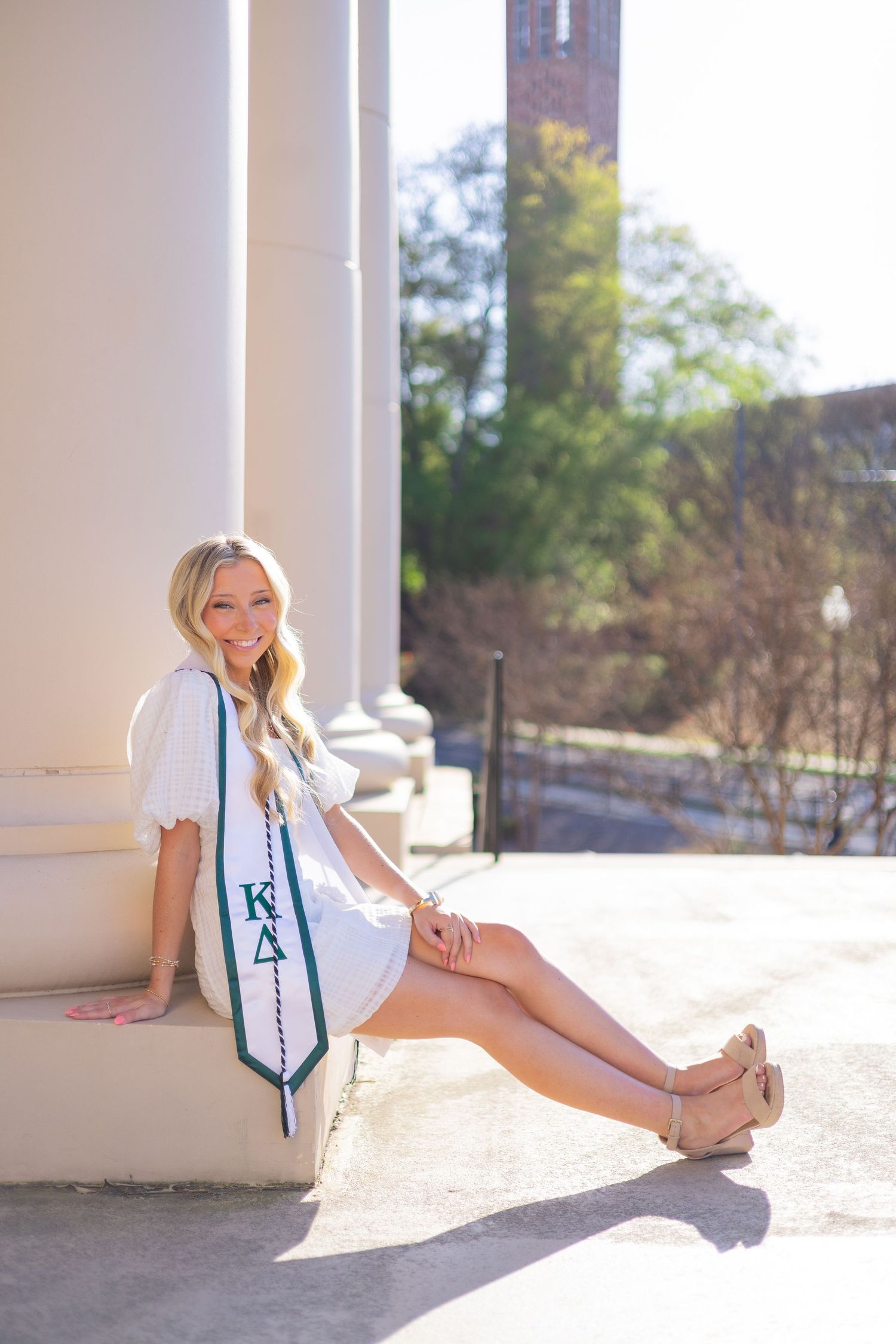 Smiling blonde college graduate in white dress wearing KD sorority stole, seated by white columns on sunny campus.