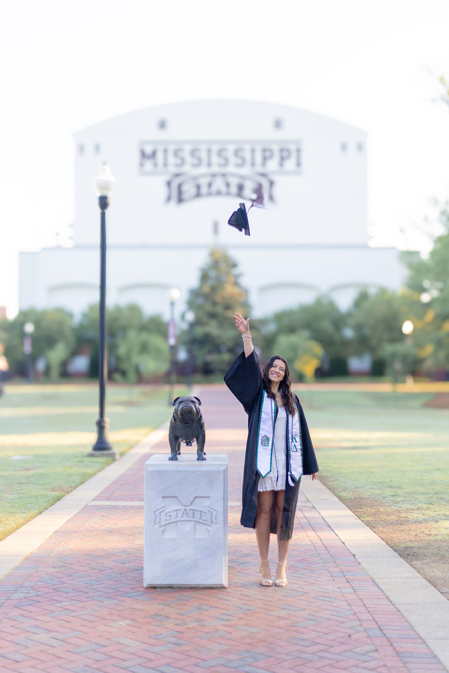Graduate throws cap in celebration next to bulldog statue at Mississippi State University.