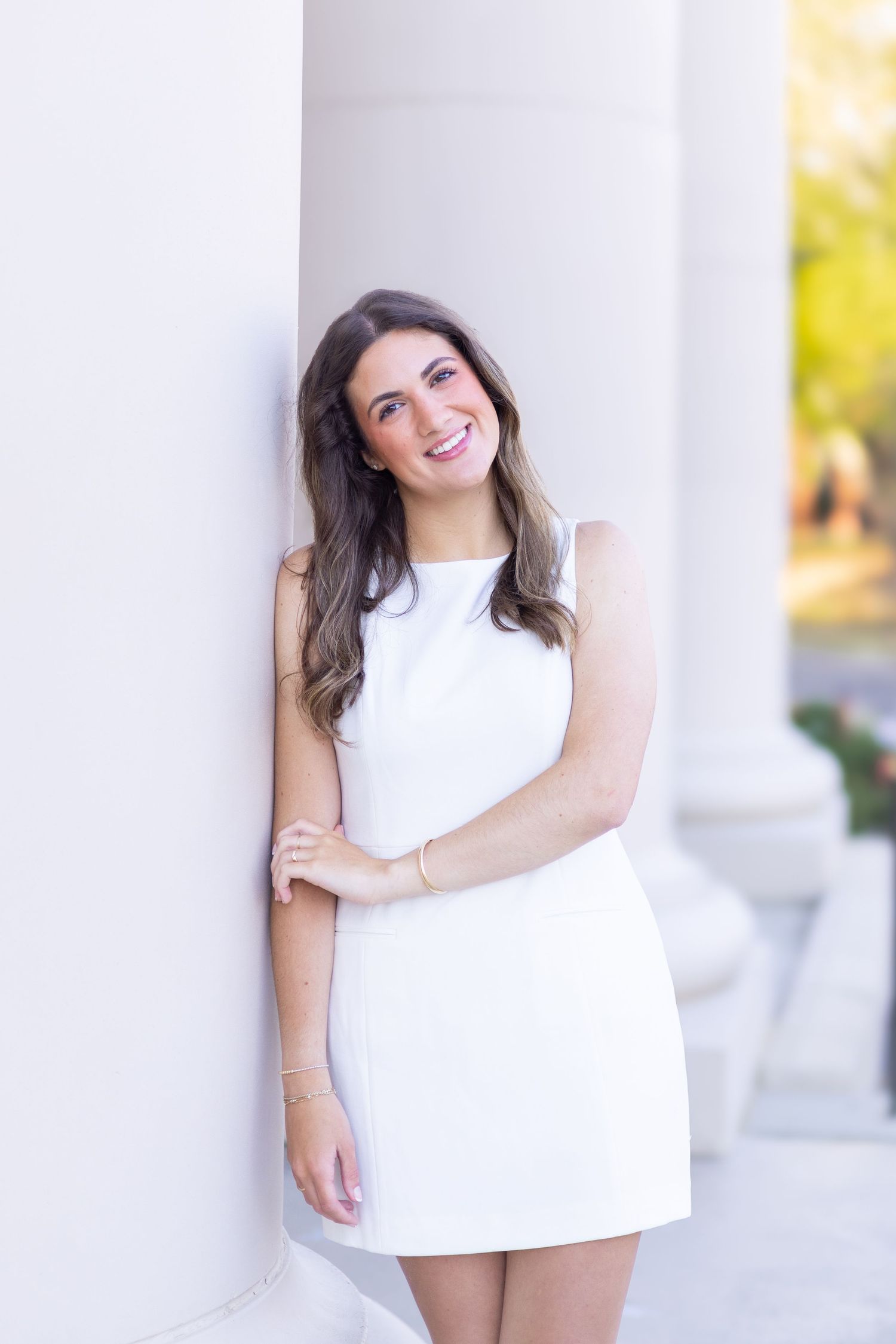 Young woman in white mini dress smiling and leaning against white columns outdoors.