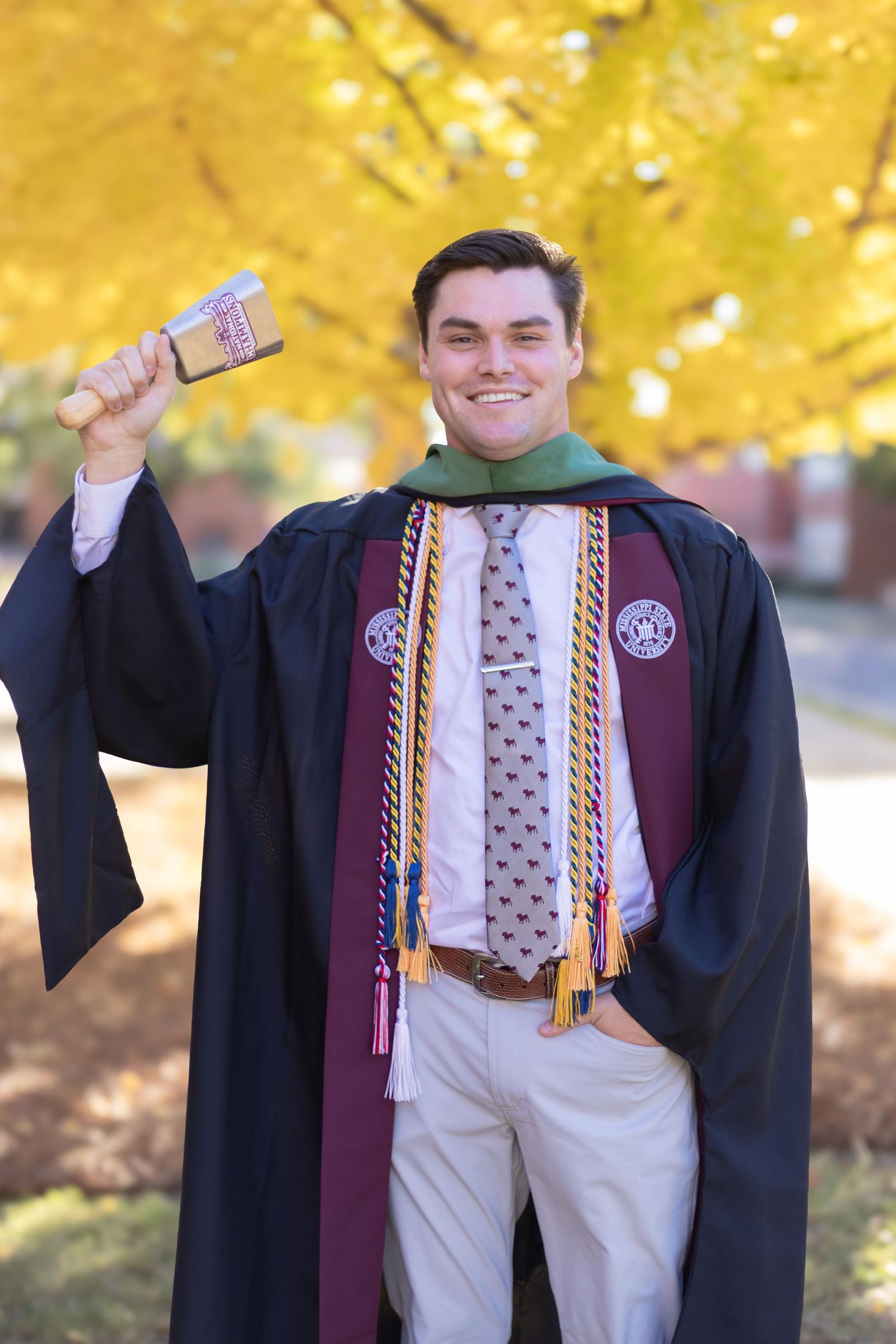 Graduate in academic regalia with honor cords poses against autumn foliage background.
