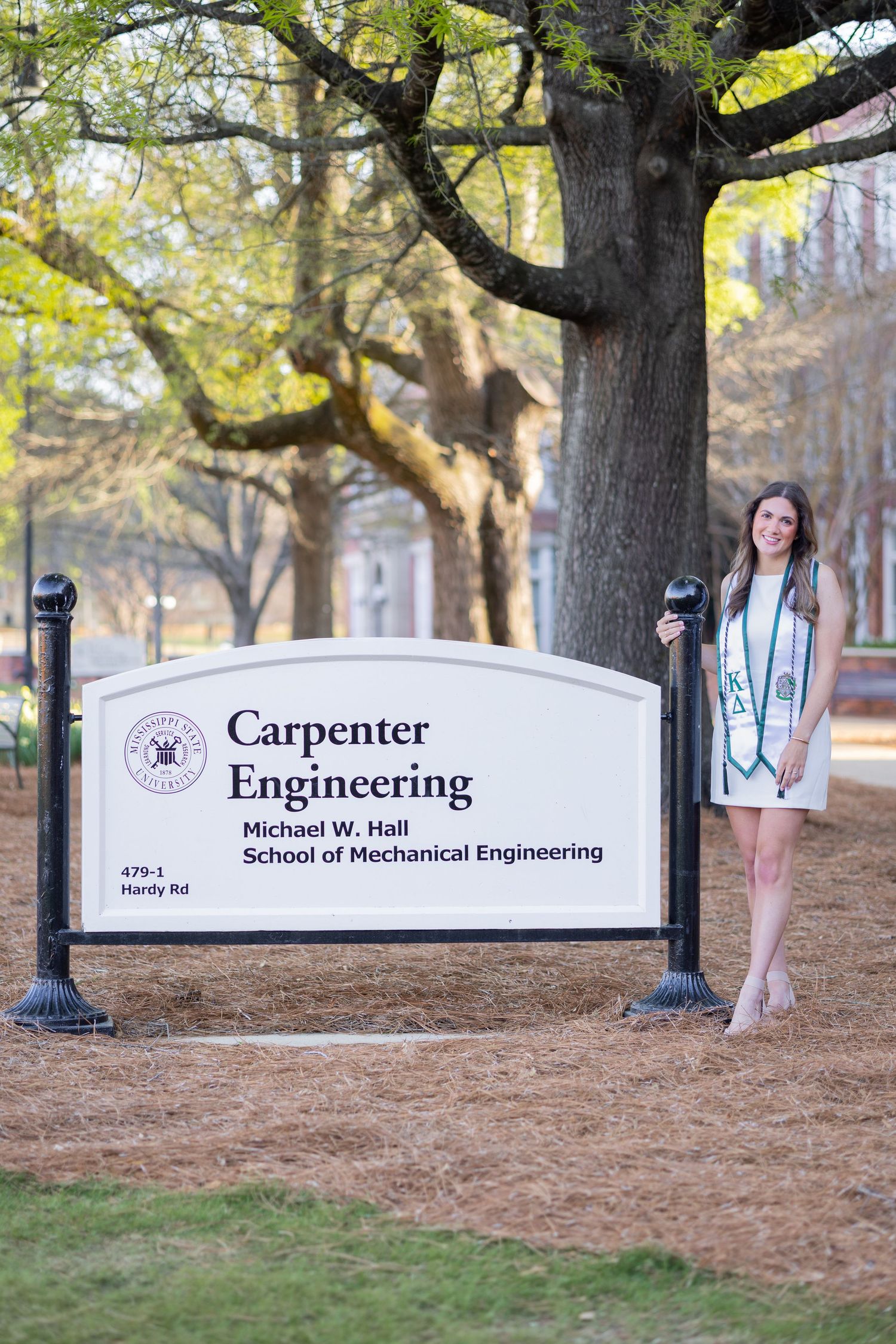 Graduate poses by Carpenter Engineering Michael W. Hall School of Mechanical Engineering sign in cap and gown.