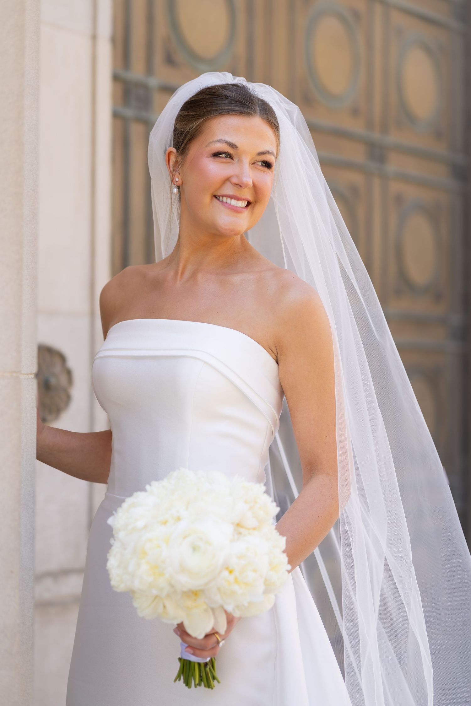 Smiling bride in strapless white gown and veil holds white rose bouquet outdoors near ornate building columns.