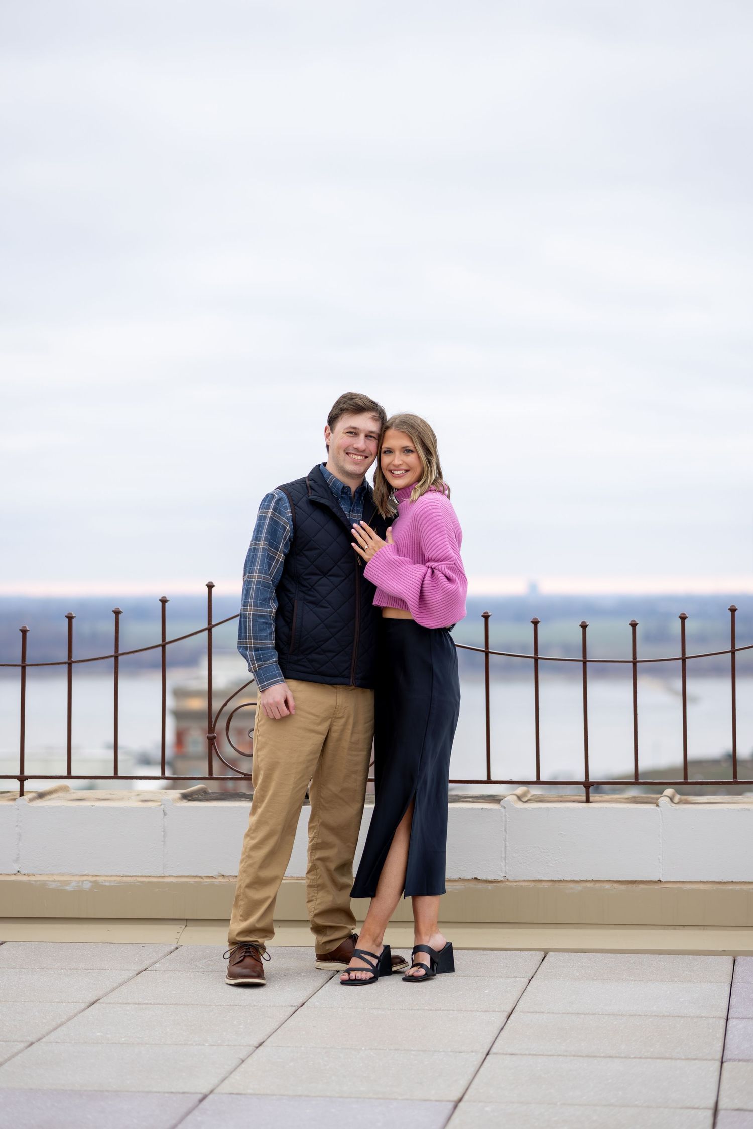Happy couple posing on a rooftop terrace, woman in pink top and navy skirt, man in plaid shirt and vest.