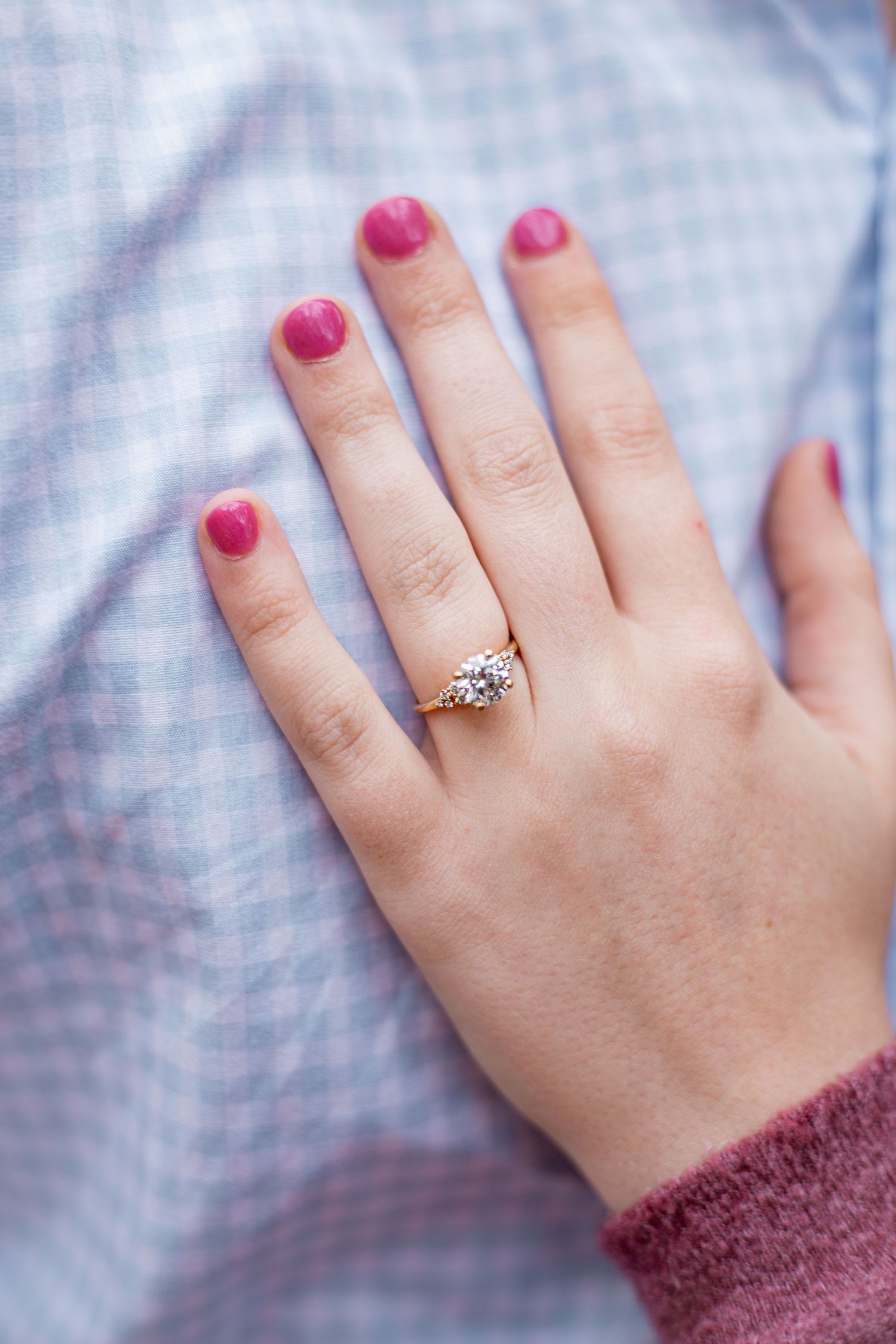 A hand wearing a rose gold engagement ring with pink nail polish against a light blue background.