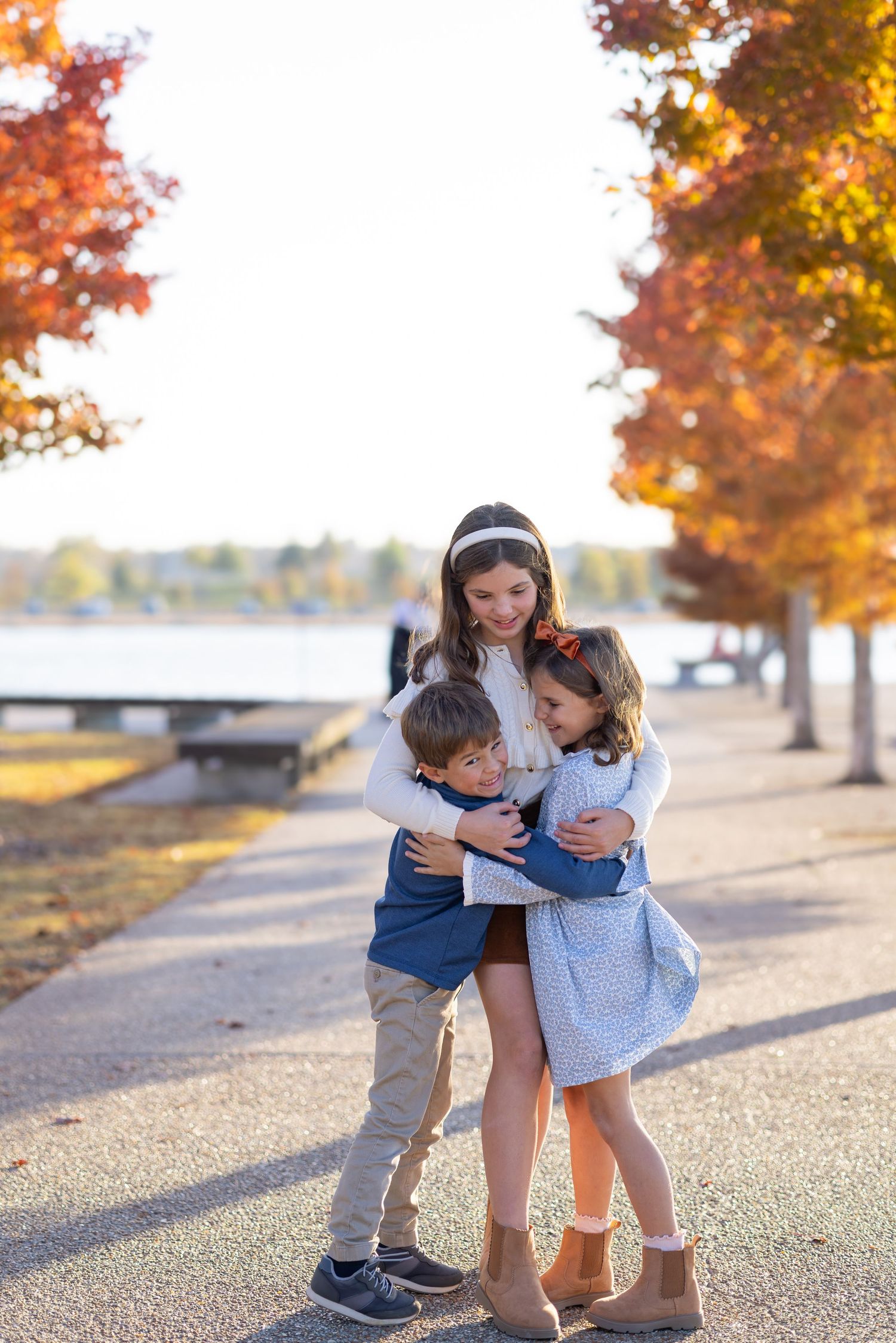 Three children share a warm hug on a tree-lined path with vibrant autumn foliage and a lake in the background.