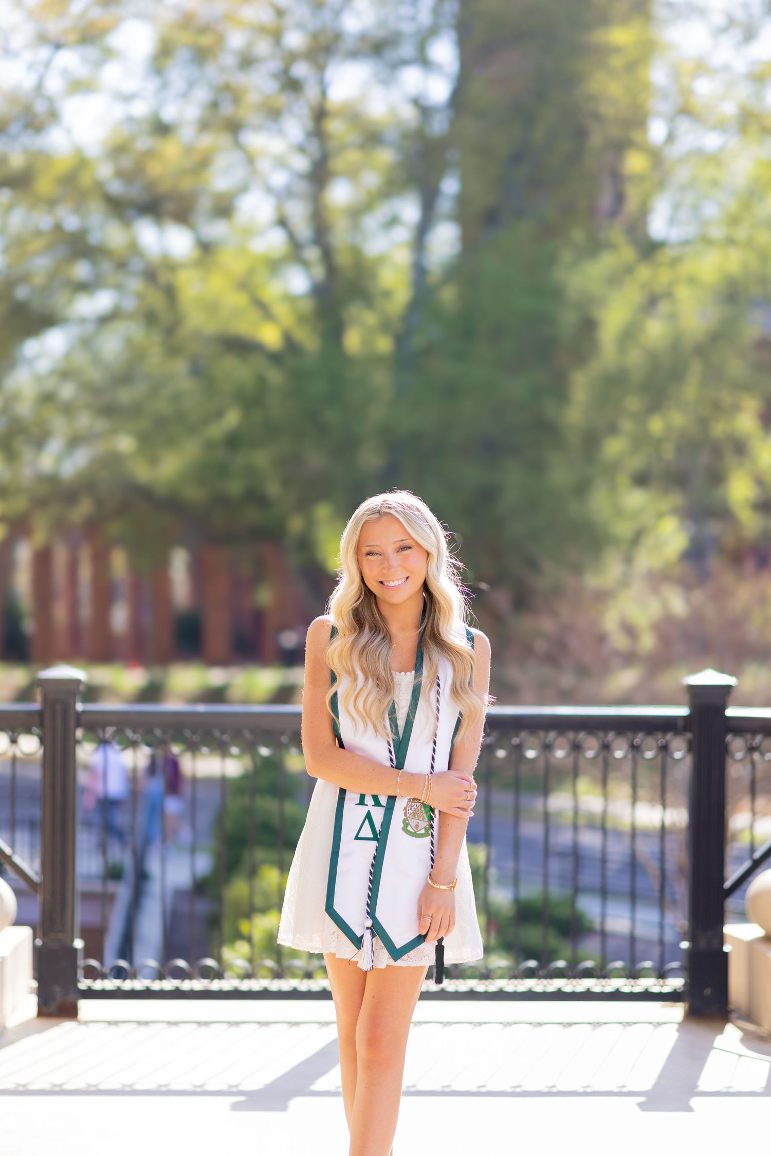 Smiling blonde woman in white dress holding graduation stole outdoors on sunny campus bridge.