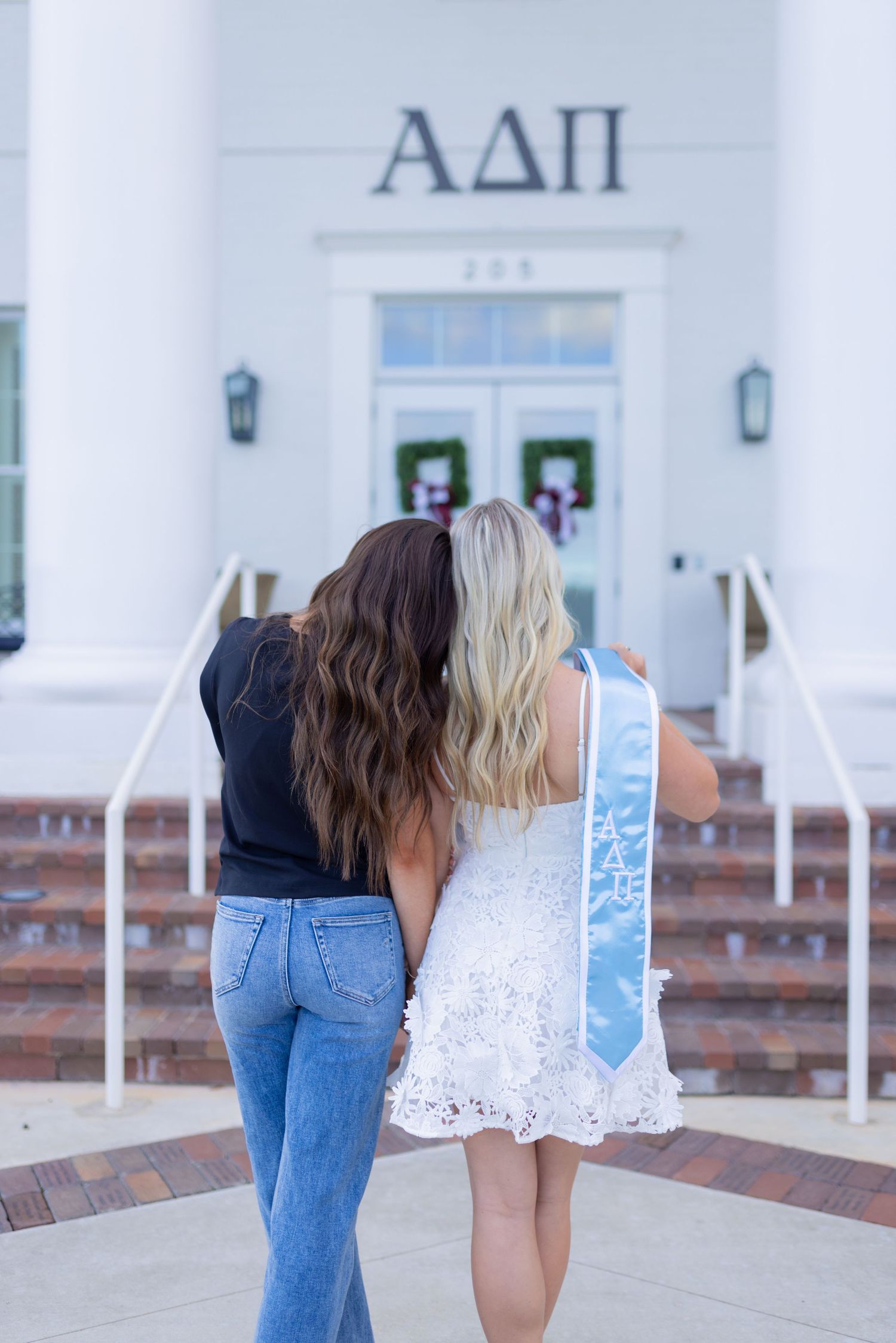 Two graduates in white dress and jeans lean forward together in front of a sorority house.