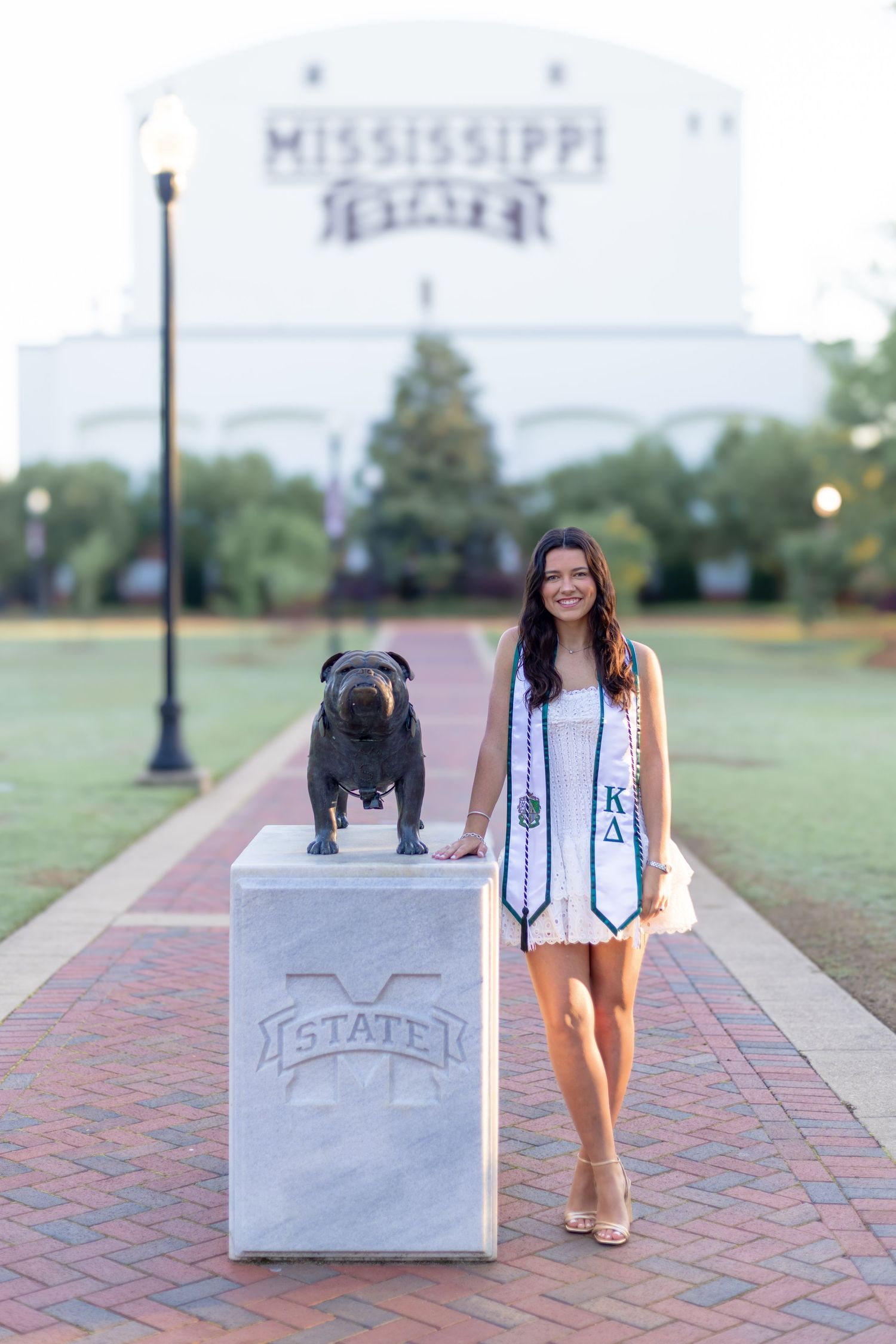 Graduate in white dress poses with bulldog statue at Mississippi State University campus.