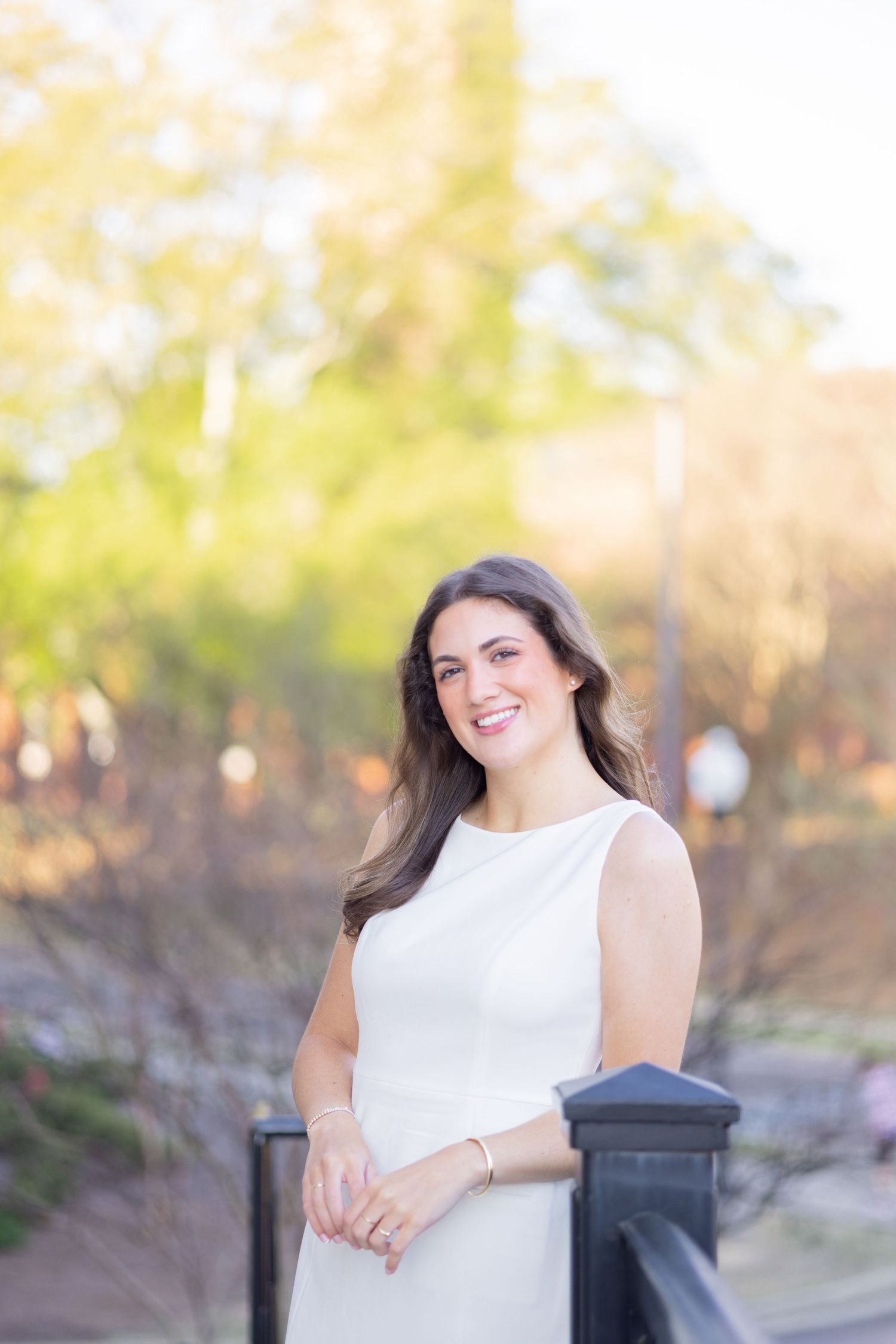 Smiling young woman in white sleeveless dress standing by black fence in sunny outdoor park setting.